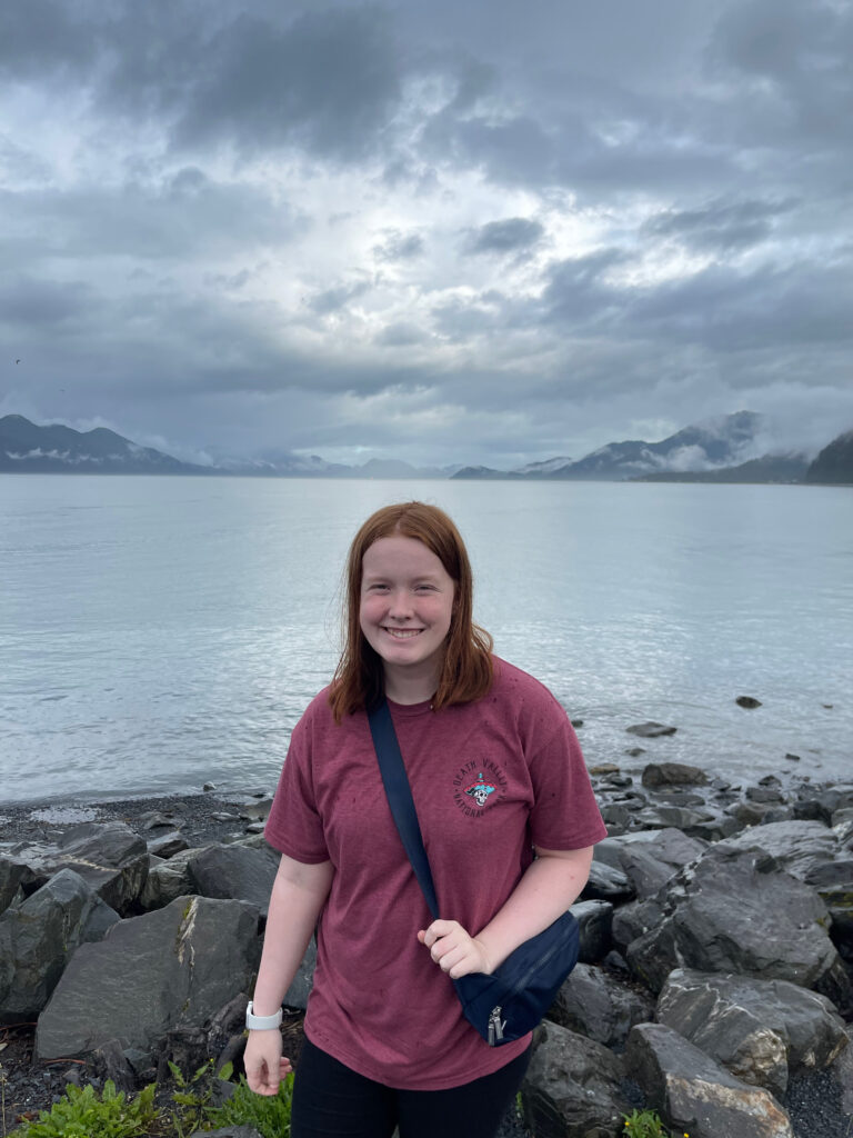 Cameron wearing a red t-shirt standing on the rocks at the edge of the bay, in Hoben Park in Seward Alaska. 