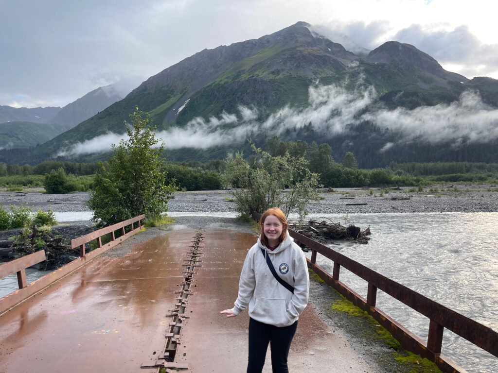 Cameron wearing a sweatshirt standing on what was an old bridge that now ends in the middle of the Resurrection River. It has just stopped raining and whats left of the metal bridge is wet. Low clouds float above the ground but below the mountain tops in the background. 