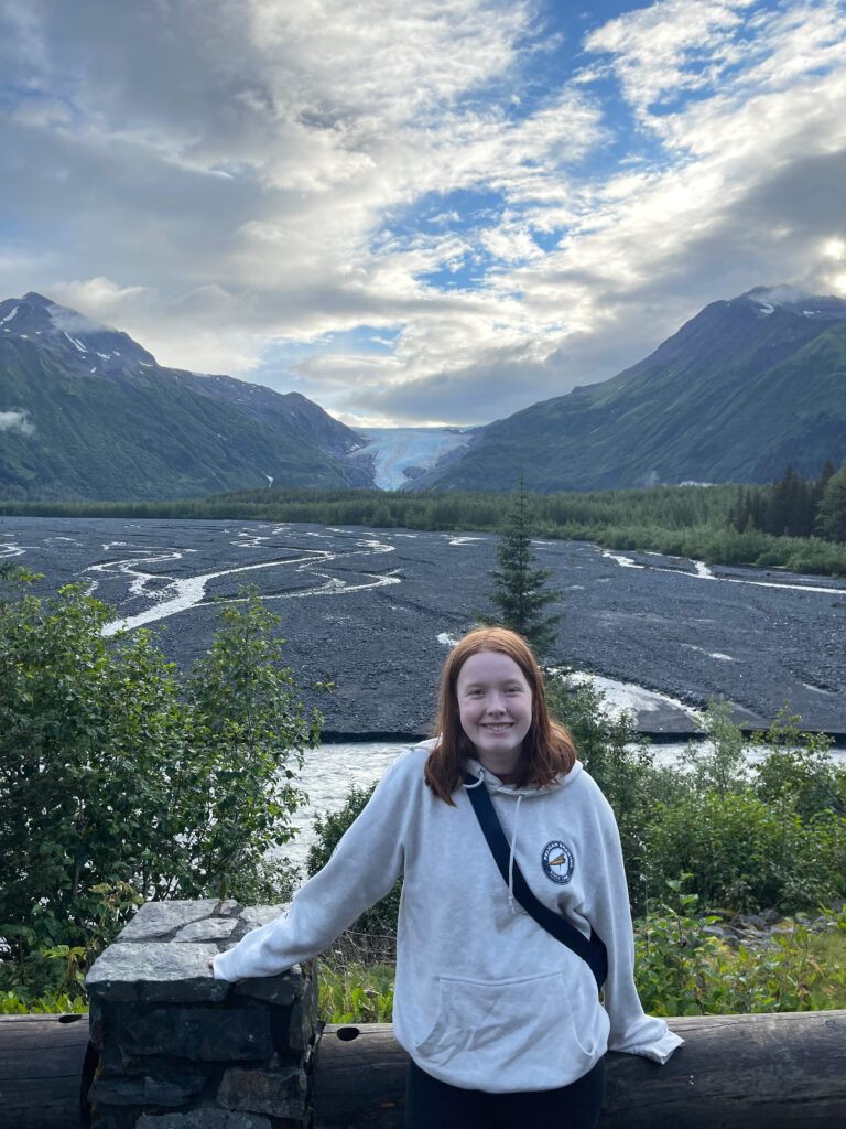 Cameron wearing a sweatshirt and standing next to the wall at Exit Glacier overlook. The massive glacier, along with stunning Alaskan mountains are in the background. Just enough light is making it way through the clouds to light up her red hair.