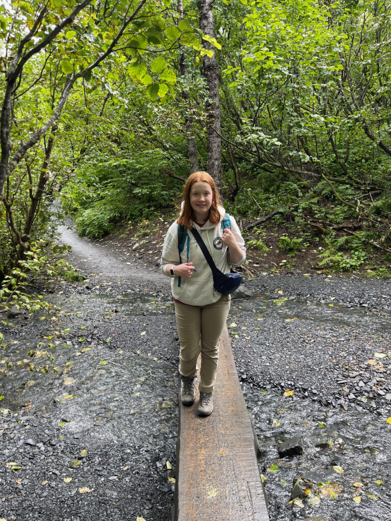 Cameron hiking deep in the wood, walking over a wooden board that goes over a mostly dry creek bed on the Exit Glacier Trail.