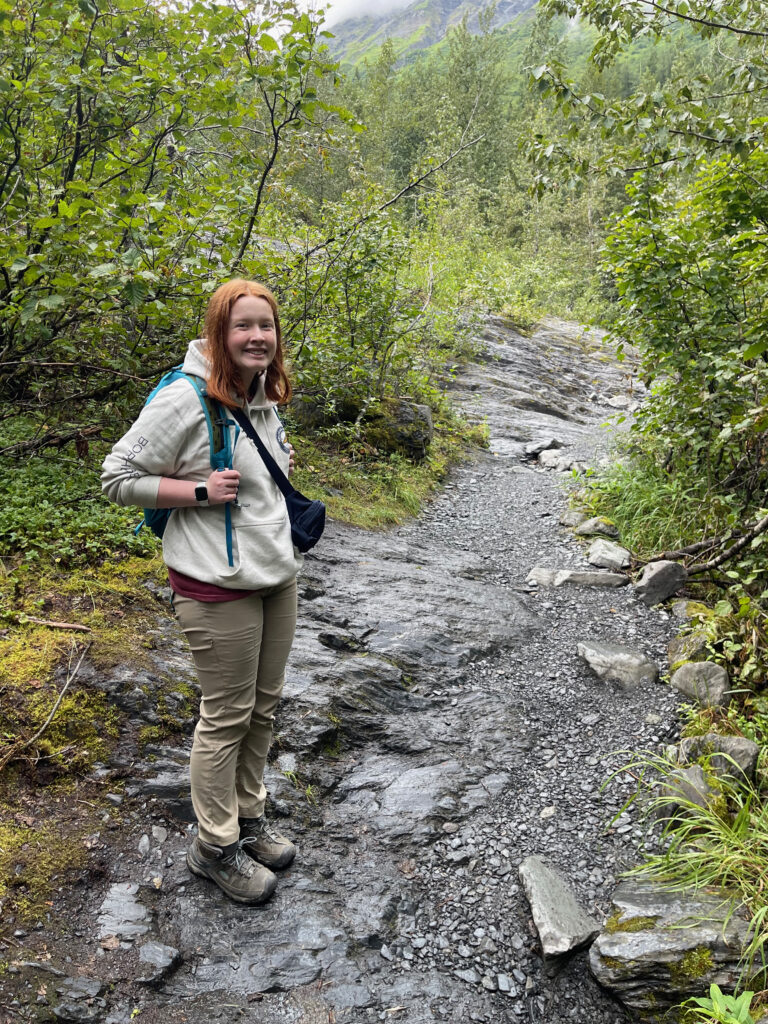 Cameron wearing her hiking gear, with a backpack, hiking the exit glacier trail, standing on very wet rocks that comprise the trail.