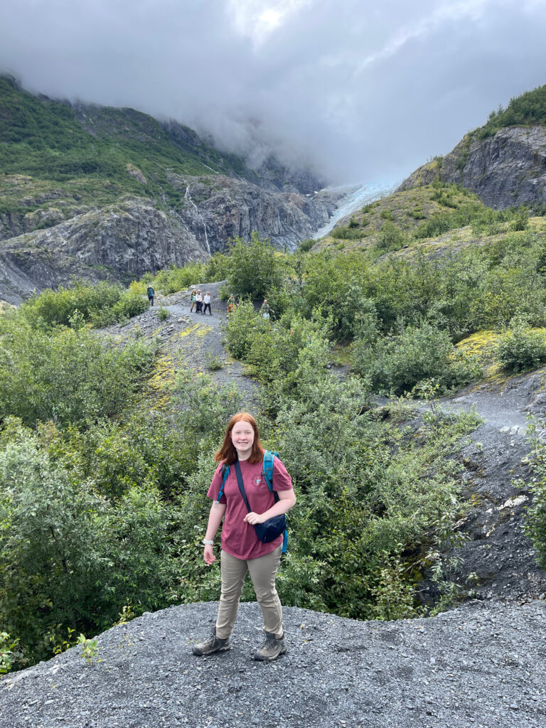 Cameron in a red tshirt and hiking pants, wearing a hiking backpack. Stopped on an open area of the Exit Glacier trial - people can be seen in the distance and beyond them the glacier itself. Clouds are coming down and covering the top of the mountain.