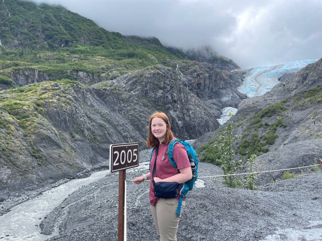 Cameron smiles at the end of the Exit Glacier Trail, next to a sign that says "2005" and marks where the glacier ended that year. In the distance you can see where the glacier is today.