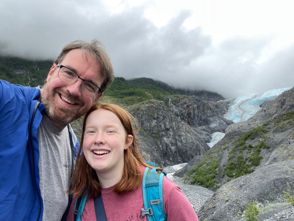 Cameron and myself, both standing and all smiles at the end of the trail. Exit Glacier and low clouds that cover the tops of the mountains can be seen in the distance. 