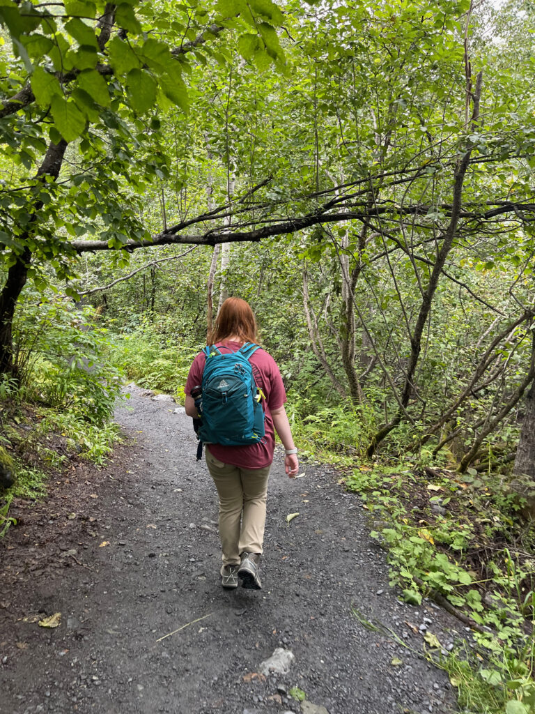 Cameron walking down the Exit Glacier Trail, wearing her red t-shirt and backpack, here the path is a bit wider and a deep very green forest from the recent rain.