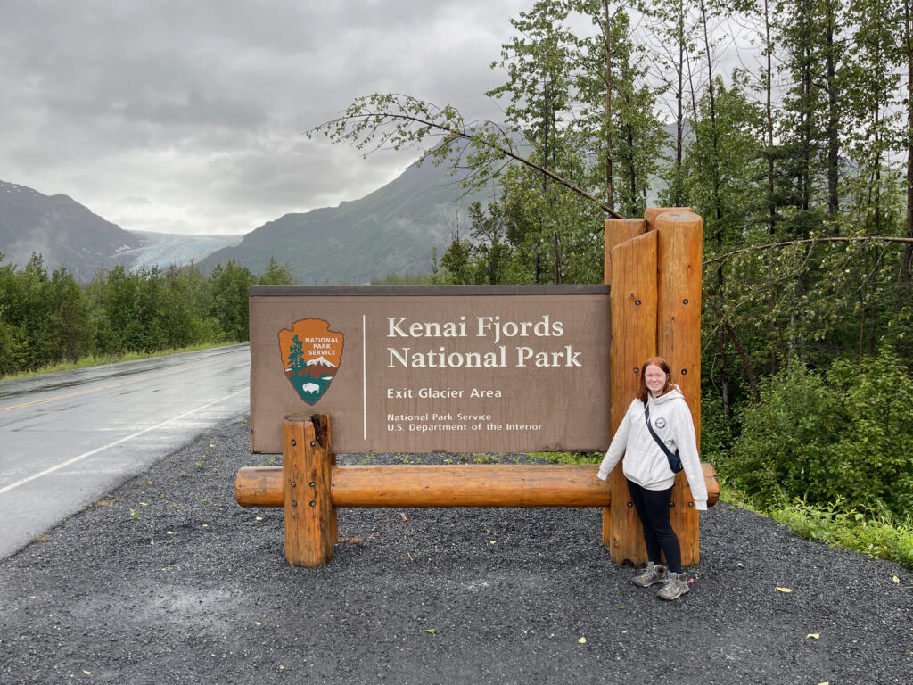 Cameron with a big smile standing in front of the sign for Kenai Fjords Natioanl Park, with the Exit Glacier and mountains in the background. 