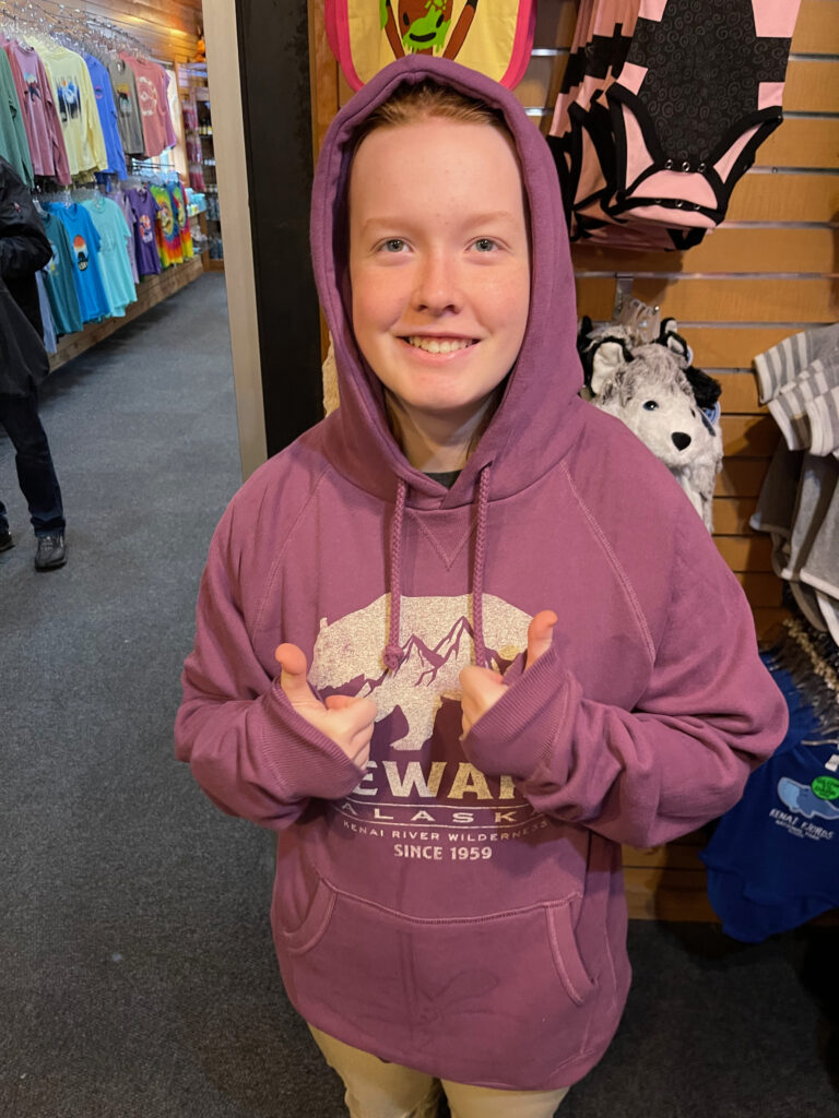 Cameron giving top thumbs up as she tries on a Kenai Fjords red sweatshirt in the Kenai Fjords Tours Gift Shop.