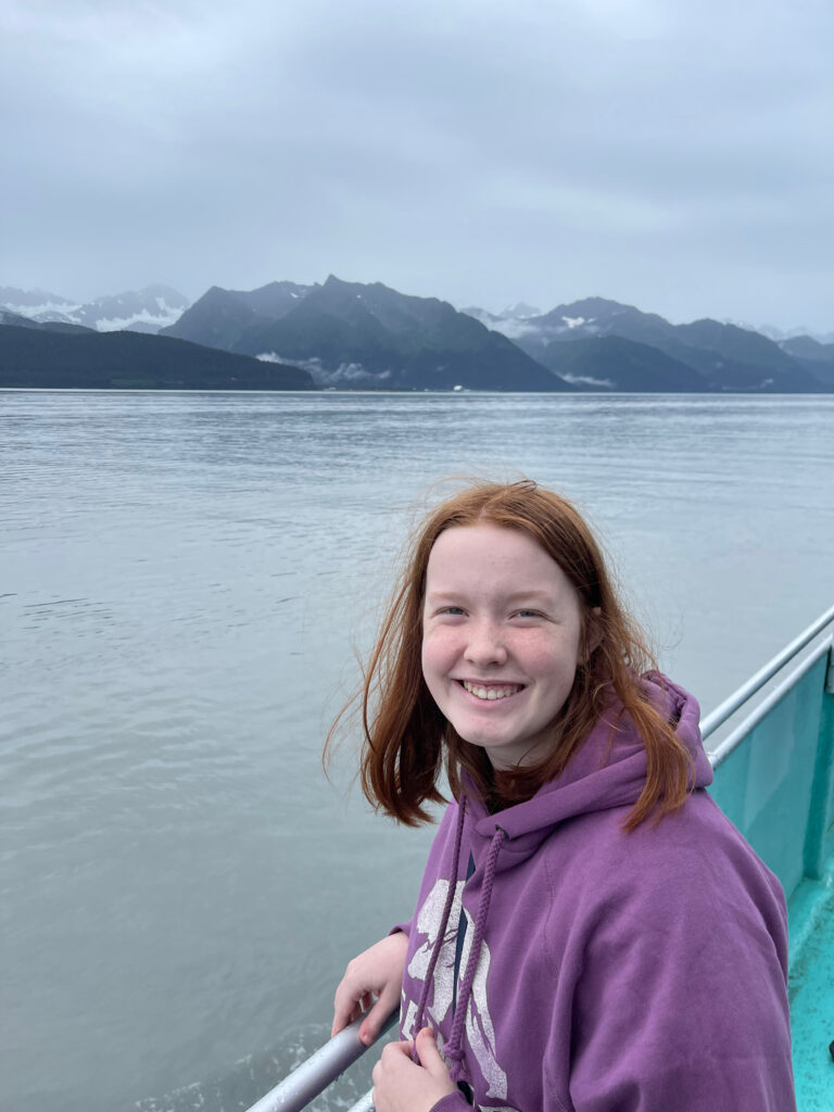 Cameron on the deck of the boat, smiling, as we make our way back to land with clouds overhead and mountains in the distance. 