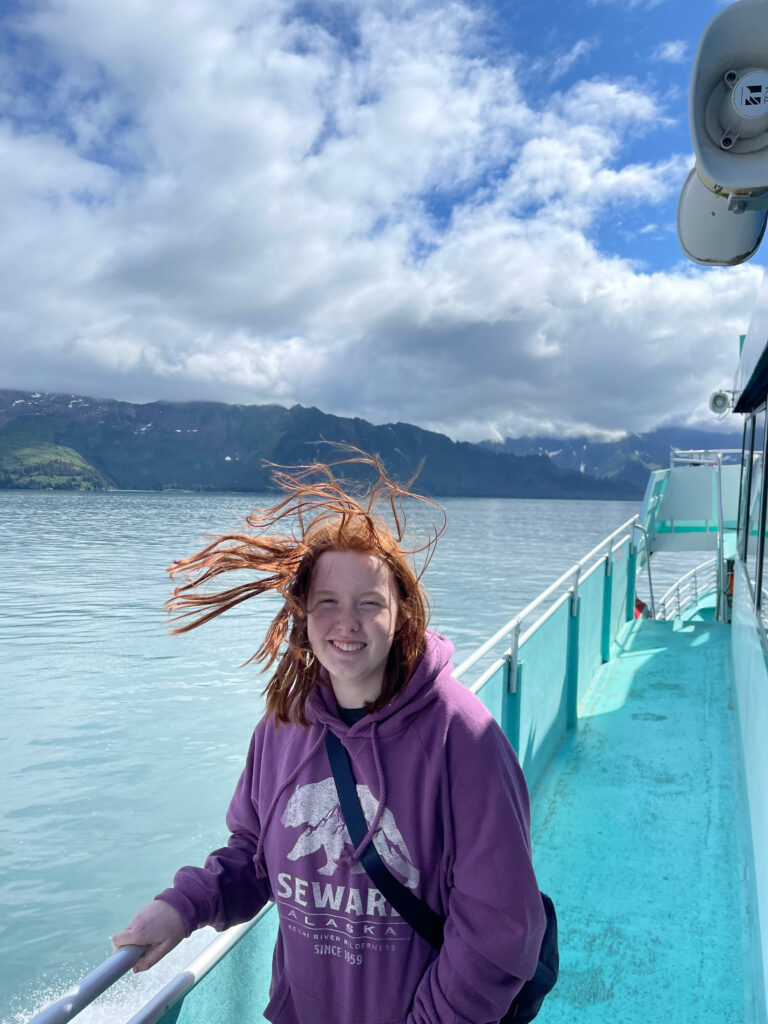 Cameron standing outside on the deck of the tour boat in Kenai Fjords National Park. The bought is a very light blue color and everyone else is inside. Her red hair is being blown all around from the wind.