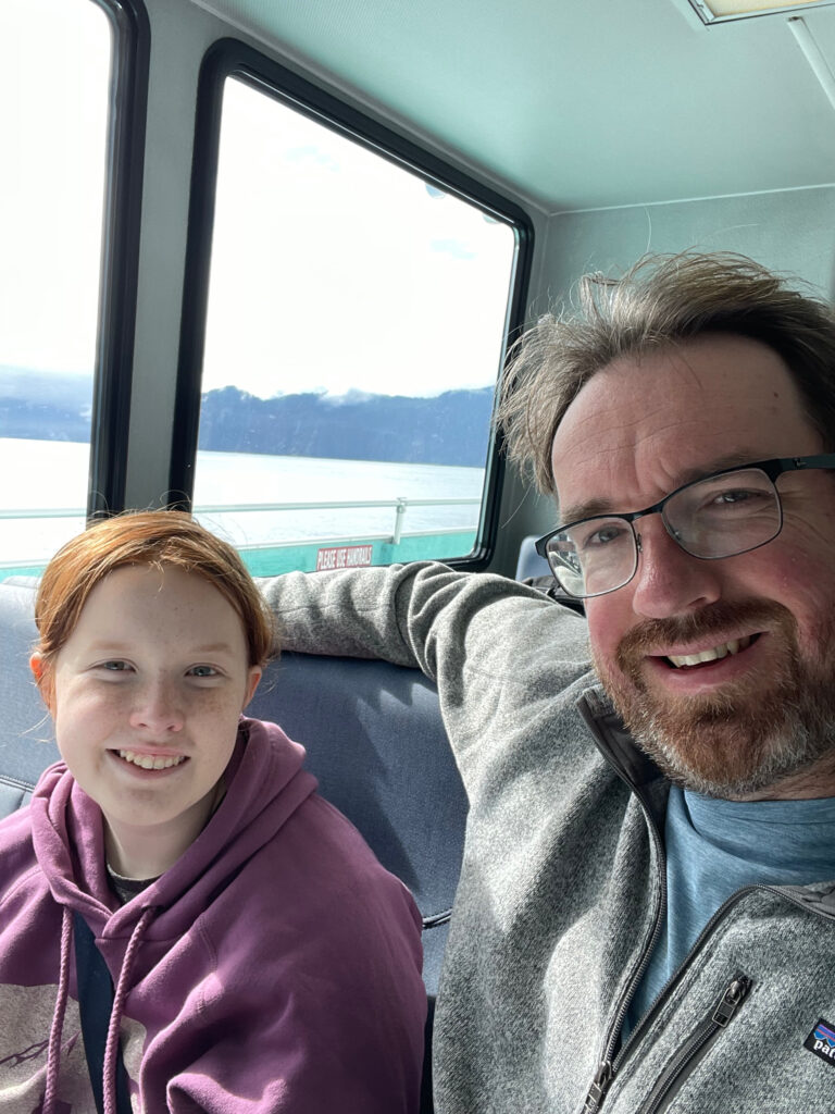 Cameron and myself sitting inside the tour boat that will take us on a tour of the coastal glaciers. We are both smiling and the large windows behind us show the view of the water and mountains. 