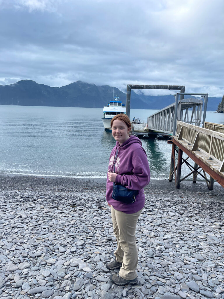 Cameron wearing a purple sweatshirt standing on the rocky Beach of Fox Island, with our boat sitting in the water at the end of the dock after just exploring all the fjords of Kenai Fjords National Park.