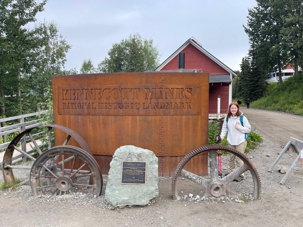 Cameron standing on the dirt road and next to the sign for Kennecott Mines National Historic Landmark, gives a thumbs up while wearing hiking clothing, a backpack and holding trekking polls. 