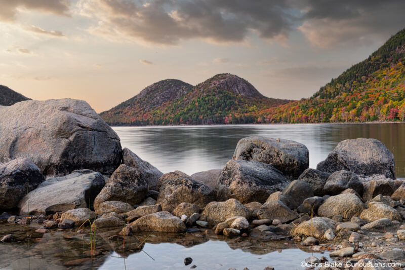 Jordan Pond at sunset in Acadia National Park with rocks in the foreground and colorful autumn mountains in the distance.