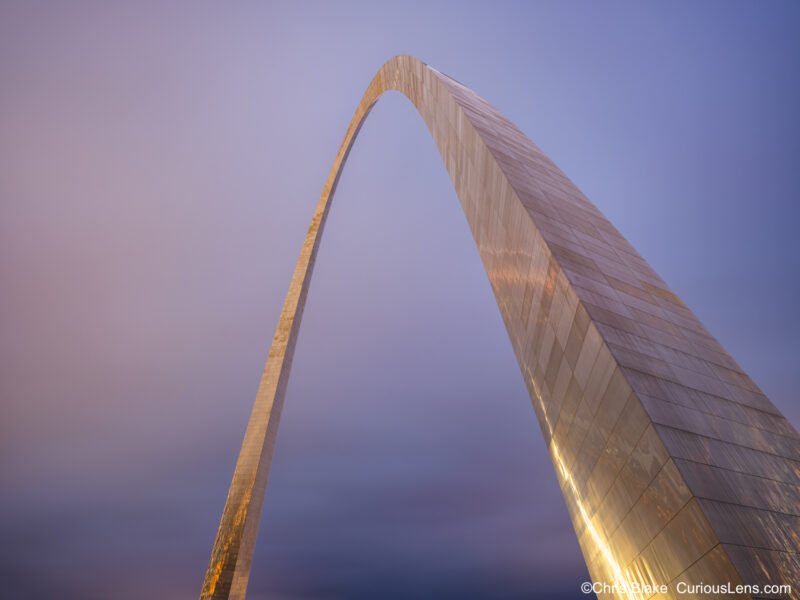 Gateway Arch in winter at blue hour with snow, freezing rain, storm clouds in warm blue and red hues, and ice-covered stainless-steel structure.