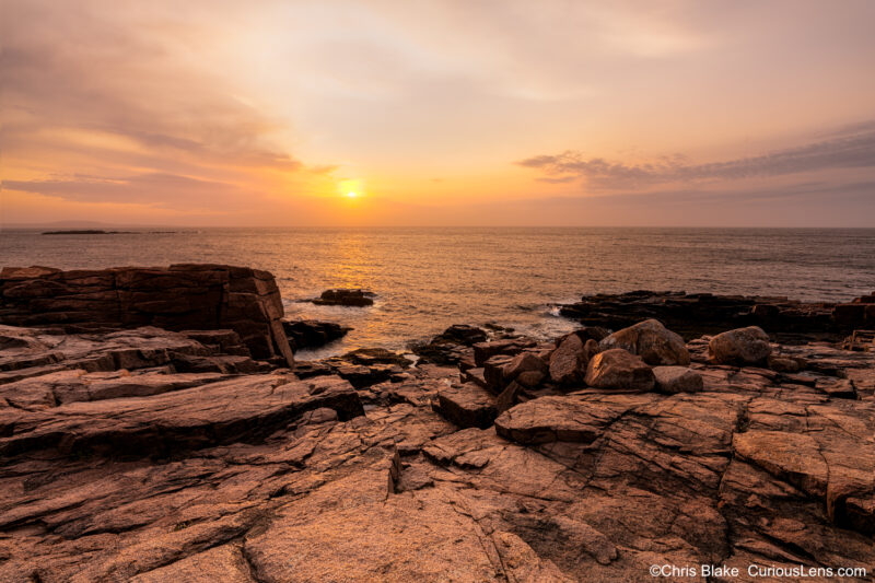 Thunder Hole at sunrise in Acadia National Park with calm ocean, colorful sky, warm red rocks, and peaceful atmosphere.