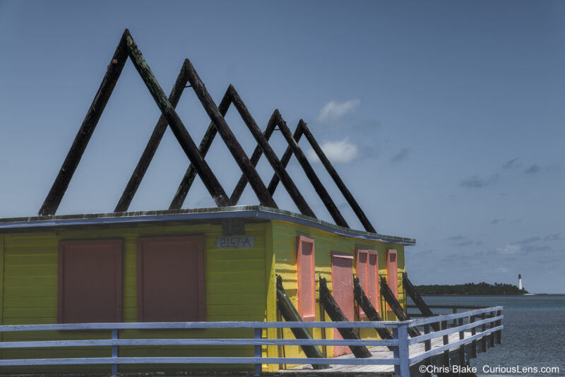 Photograph of one of the last remaining stilt houses in Stiltsville, Miami, perched above the waters of Biscayne Bay. The structure stands against a serene backdrop, with the historic Boca Chita Lighthouse visible in the distance. The image highlights the resilience of these iconic buildings, once part of a vibrant and exclusive community, now reduced to just a few surviving structures after recent hurricanes.