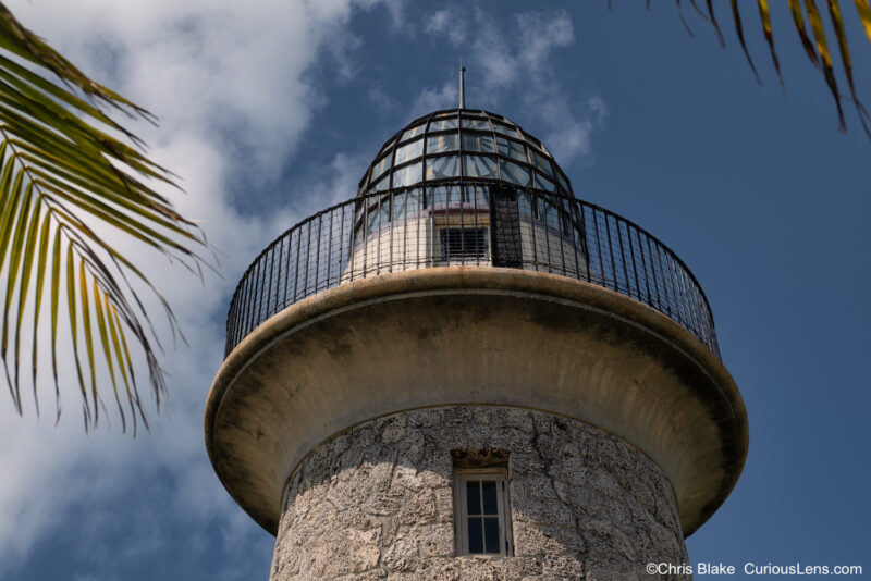 Close-up photograph of the upper level of the Boca Chita Lighthouse in Biscayne Bay National Park. The image highlights the detailed craftsmanship of the native coral rock used to build the tower, along with the glass dome roof. This iconic 1930s structure, built by Mark Honeywell, offers stunning views of nearby islands and the Miami skyline from its observation deck.