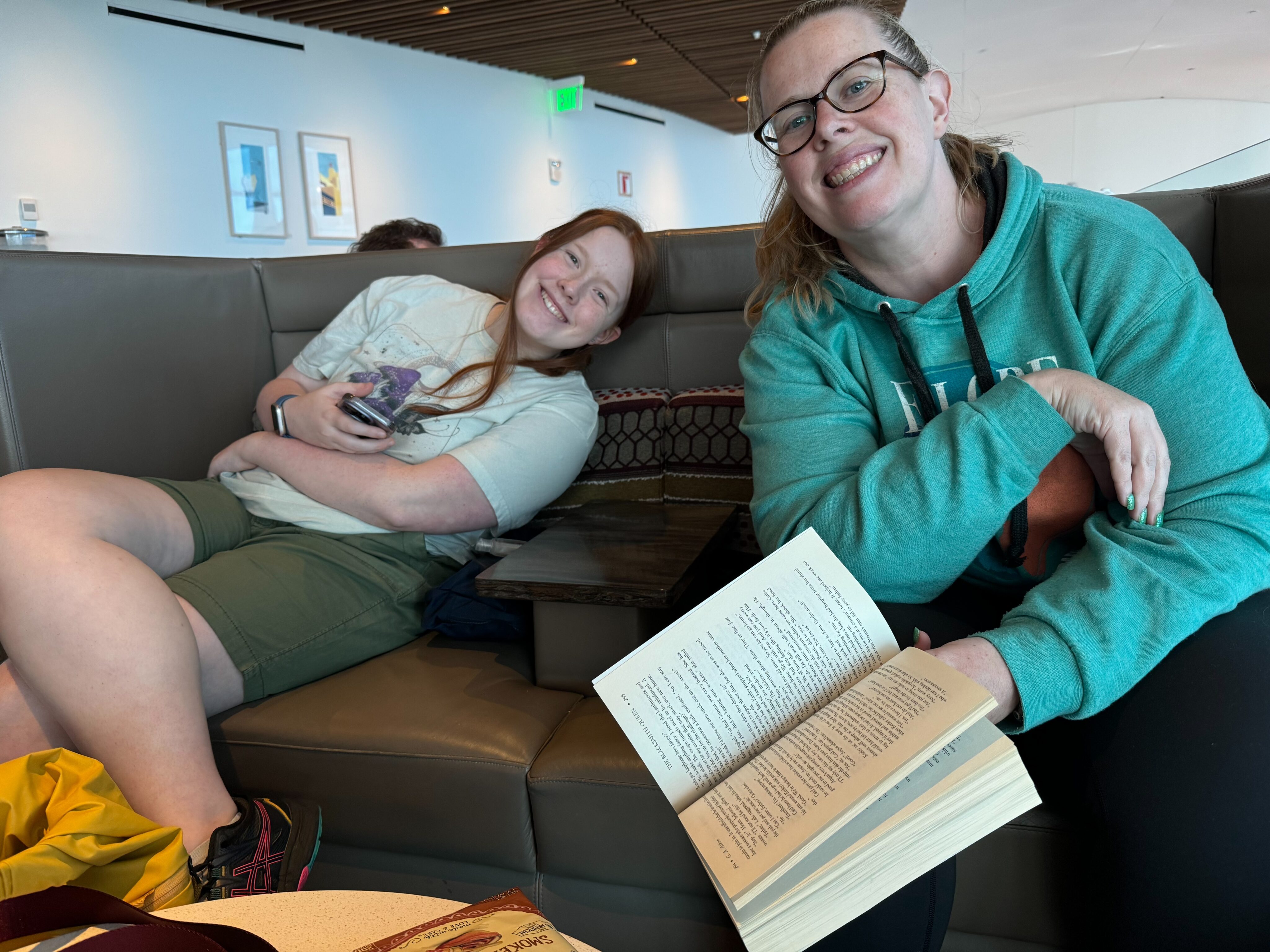 Cat holding a book, and Cami sitting back on the sofa relaxing in the Delta lounge in Seattle.