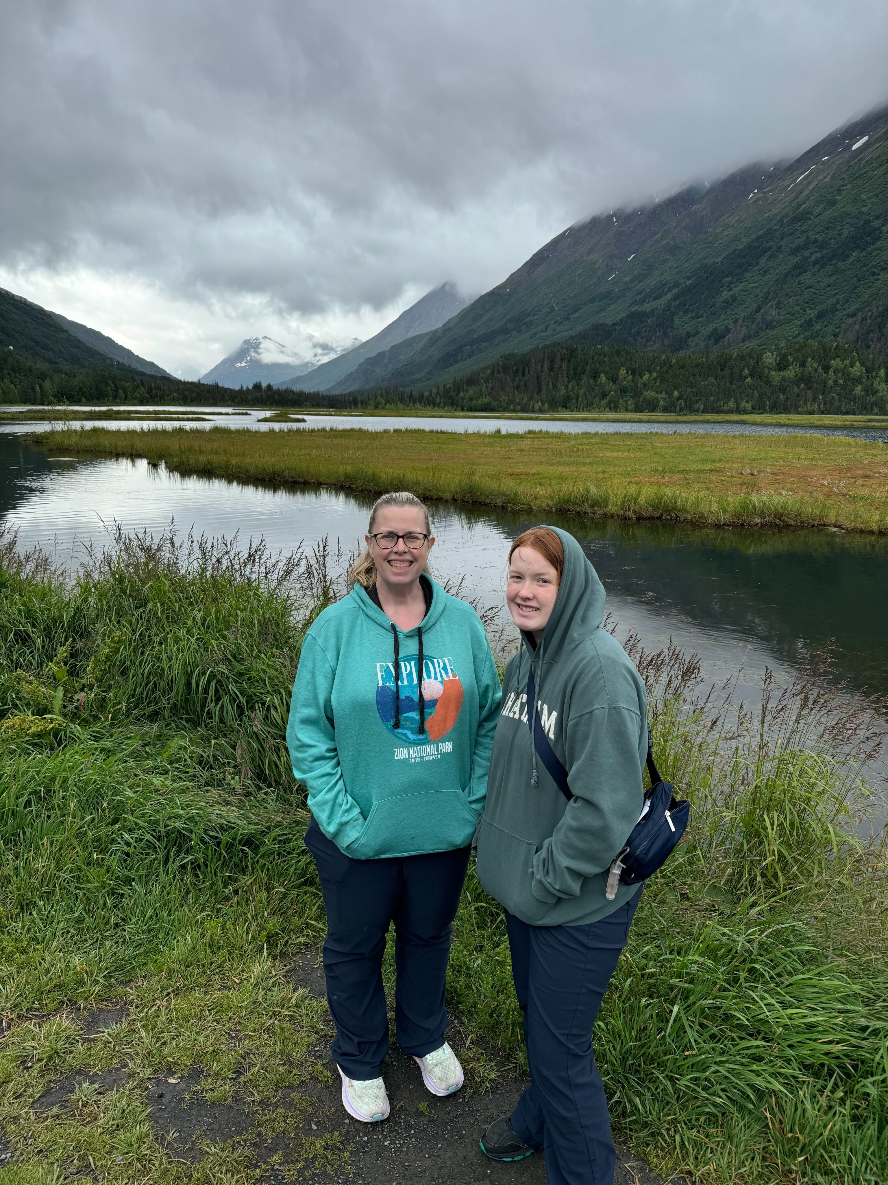 Cat and Cameron standing and both wearing hoodies at the edge of Tern Lake with clouds and mountains in the background.