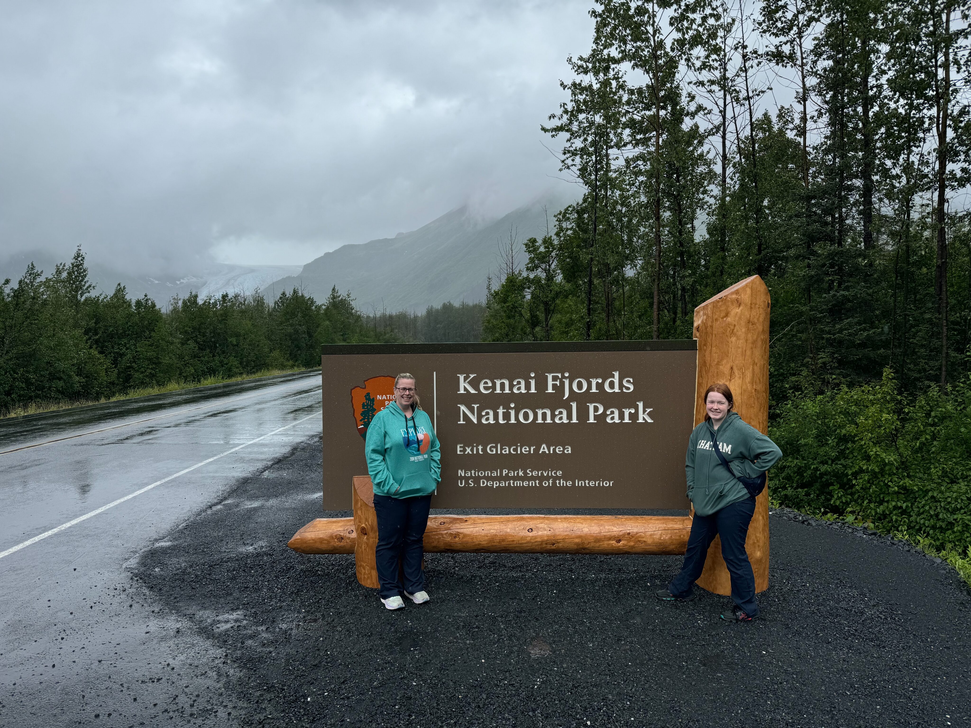 Cat and Cameron standing in front of the sign for Kenai Fjords National Park sign. On a rainy day with clouds covering most of the mountains close by. 