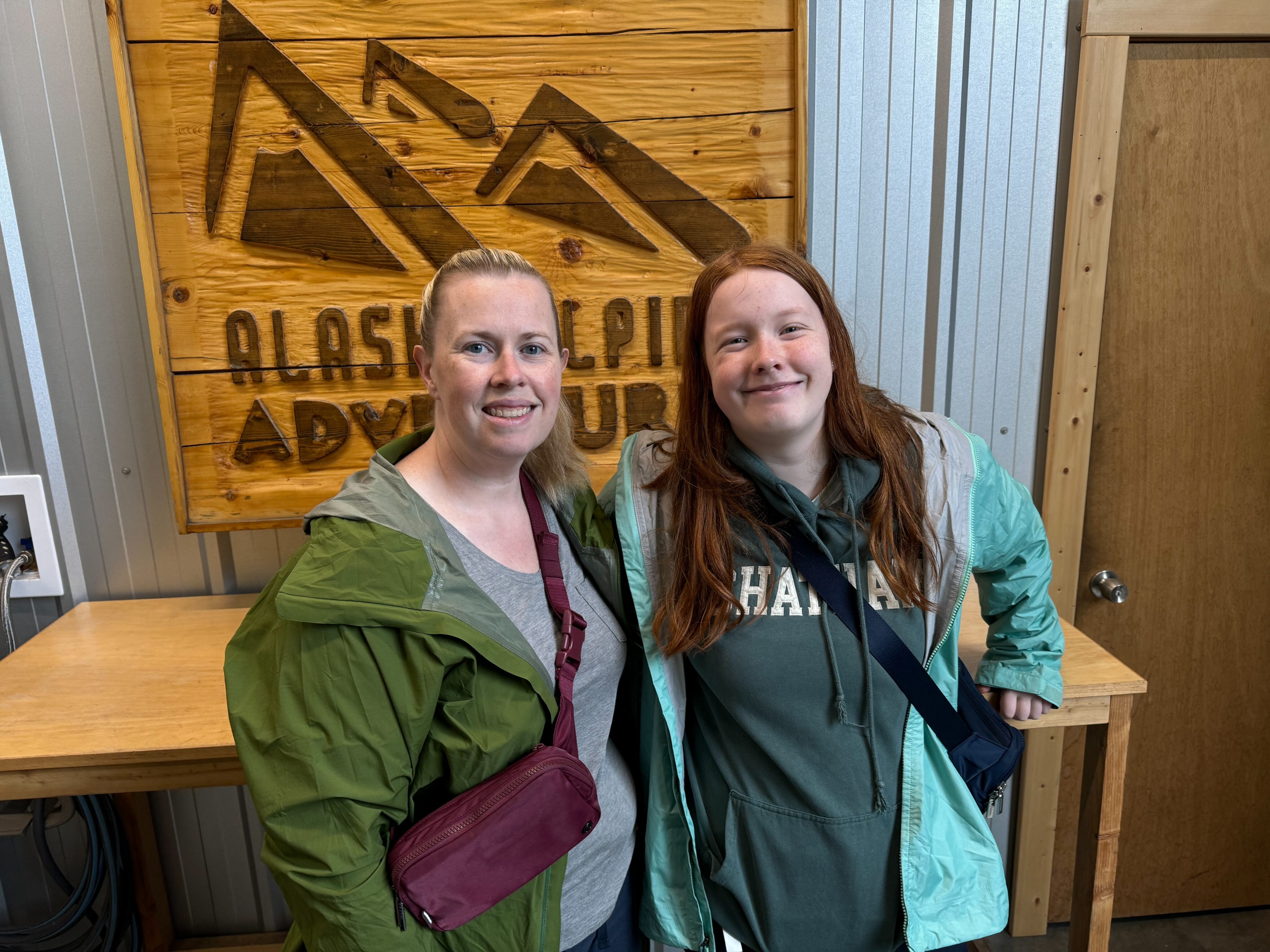 Cat and Cameron wearing raincoats and crossbody bags standing in the gear room of Alaska Alpine Adventures. 