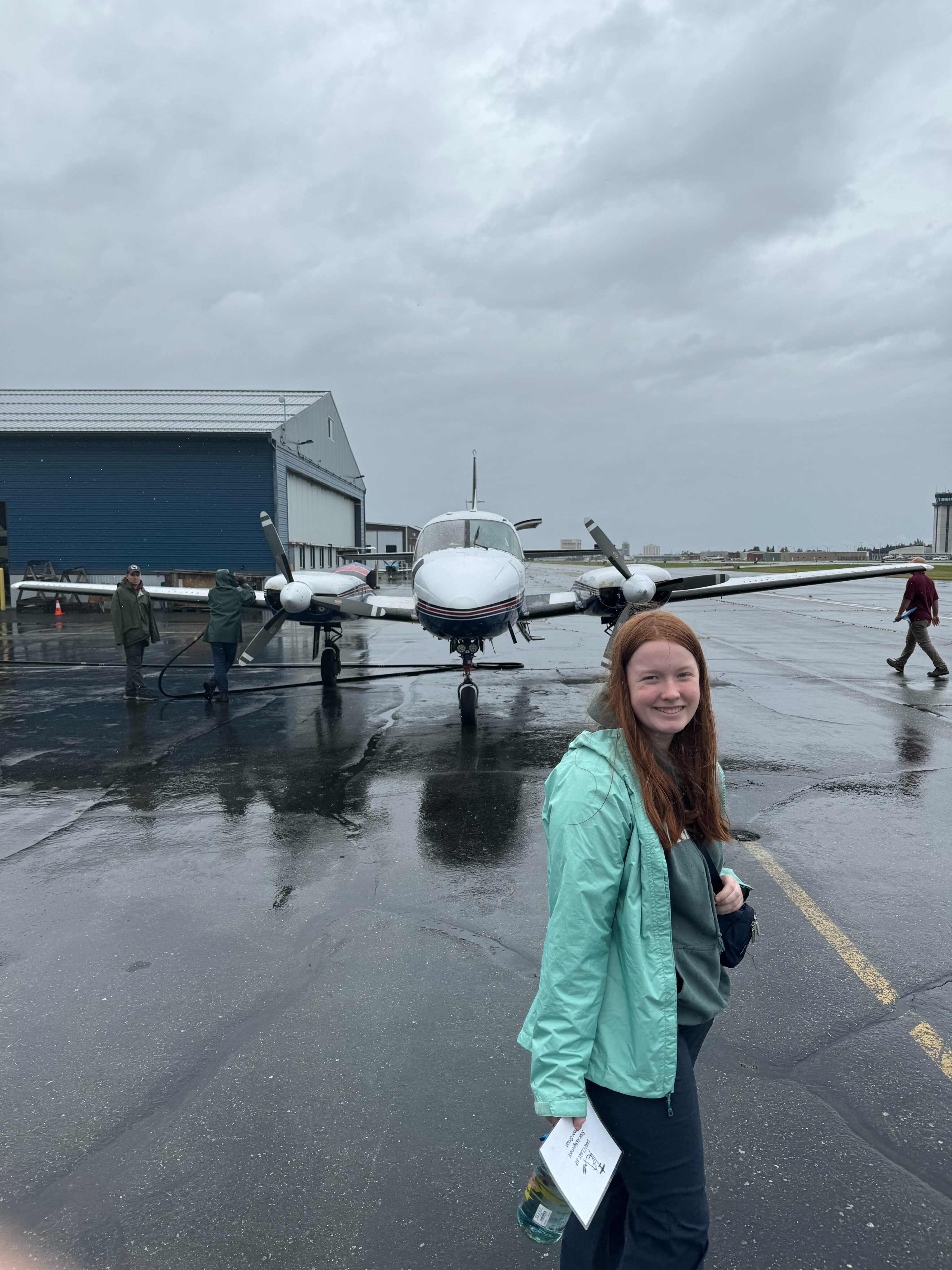 Cameron, wearing her raincoat turning around for the photo, on a rainy day on the tarmac at Merrill Field - in front of the small prop plan that Lake Clark air will use to fly us to Port Alsworth. 