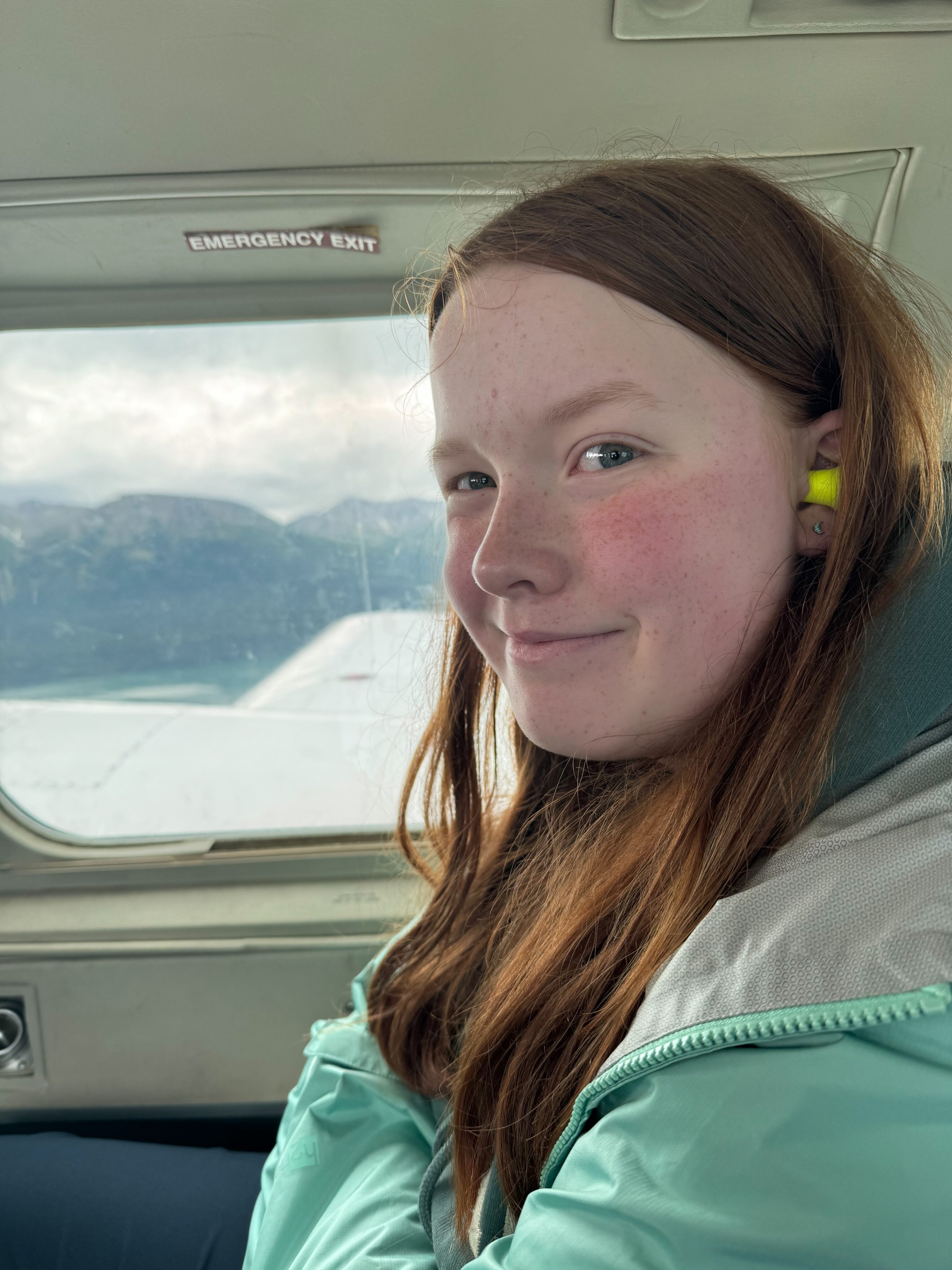 Cameron wearing her rain jacket, sitting in the window seat of a Beechcarft turboprop plane. Ear plugs are in her ear, and amazing mountains are outside her window. 