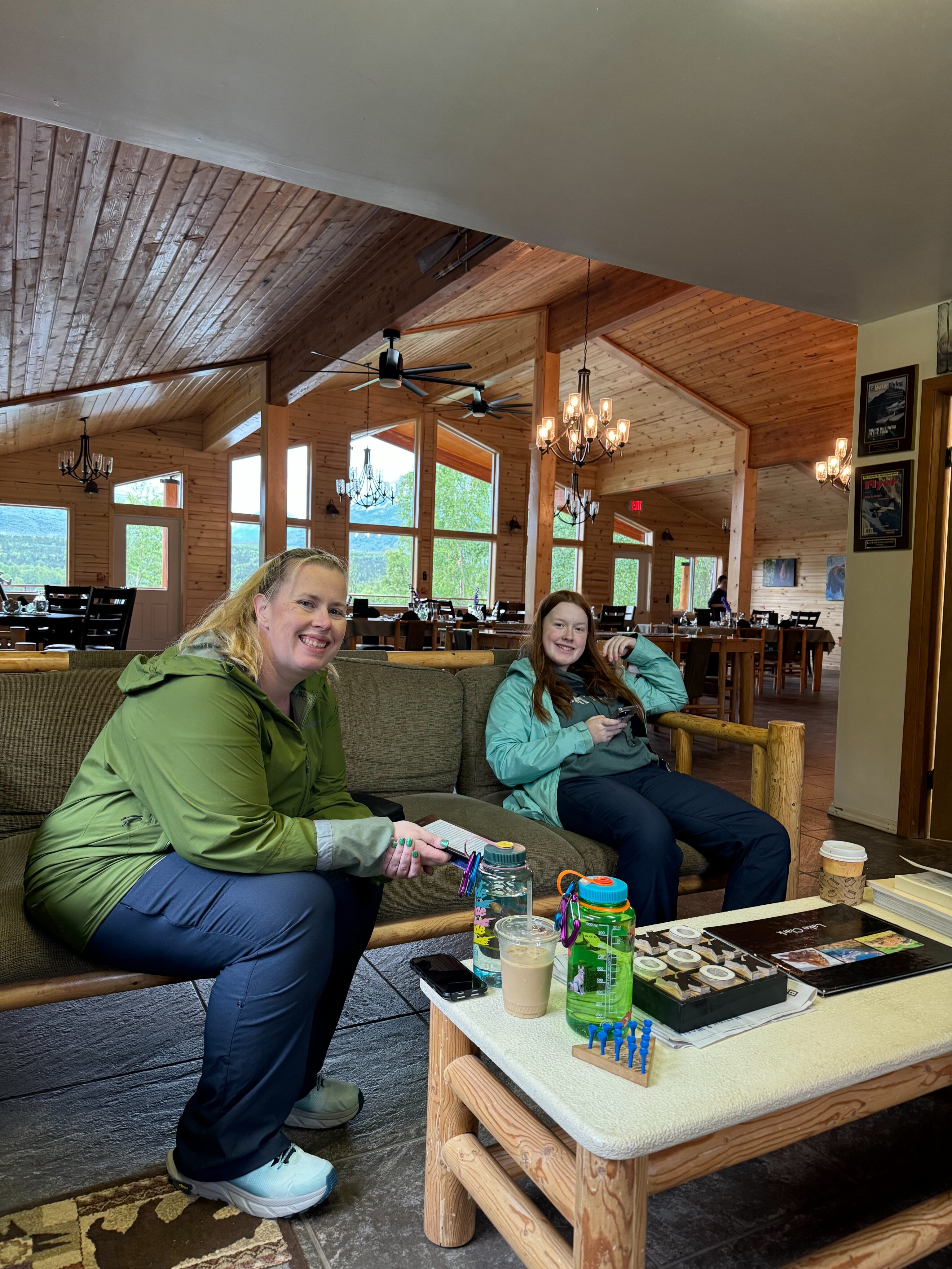 Cat and Cameron sitting on a sofa in the lodge at Port Alsworth. The dinning room and massive windows overlooking the lake are behind them.