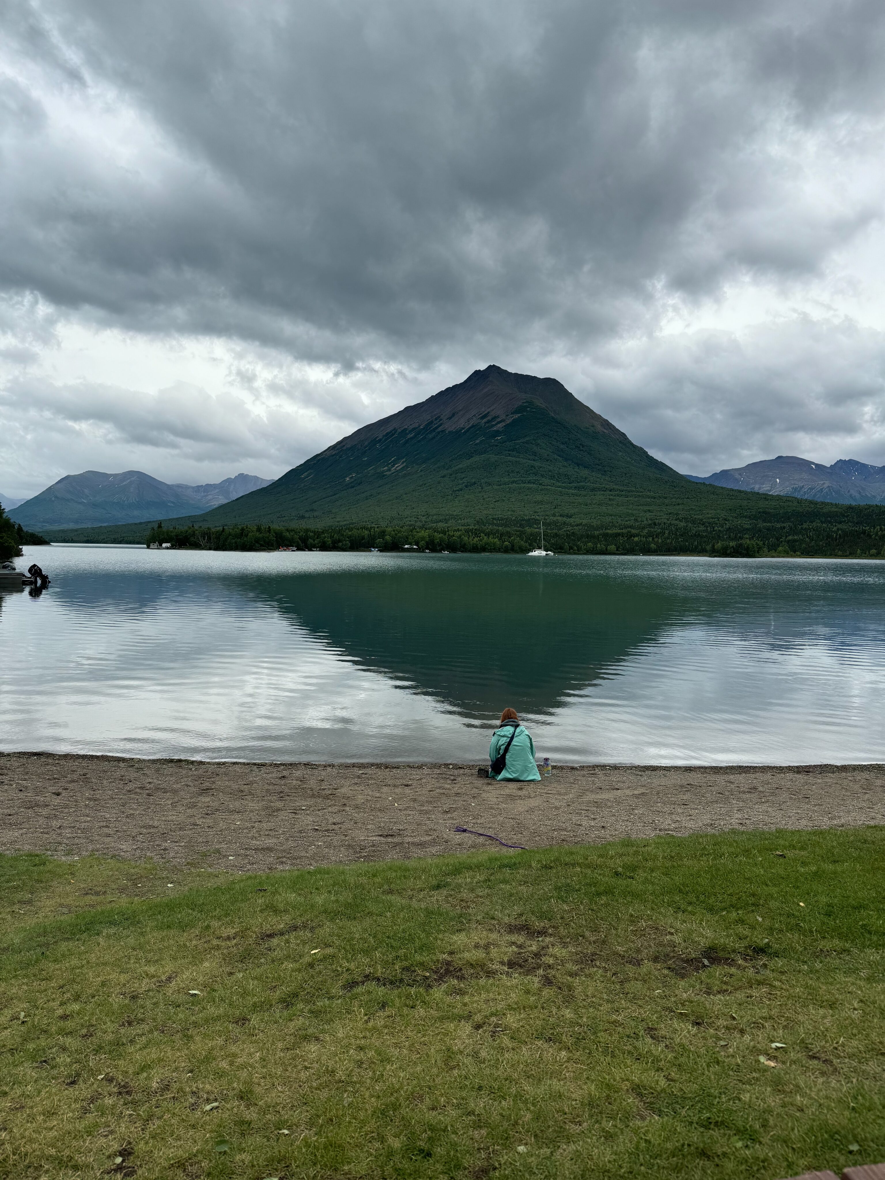 Cameron sitting by the edge of Lake Clark, with a giant mountain across from here and a perfect reflection in the water. Dark rain clouds are overhead. 