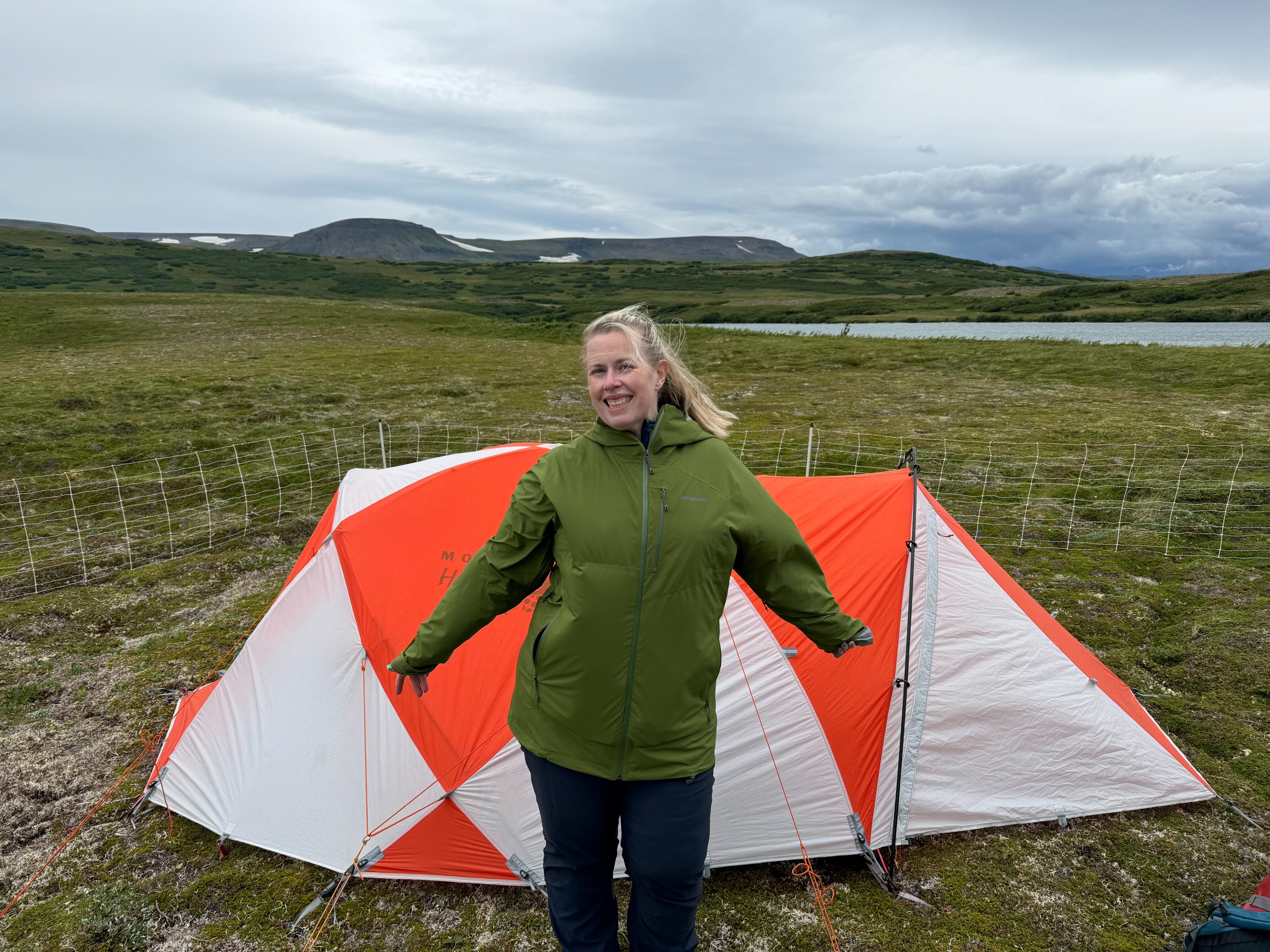 Cat in her green raincoat standing with her arms out and a big smile showing off our tent we just finished setting up. Behind us there is our electric fence, a lake and mountains in the distance.