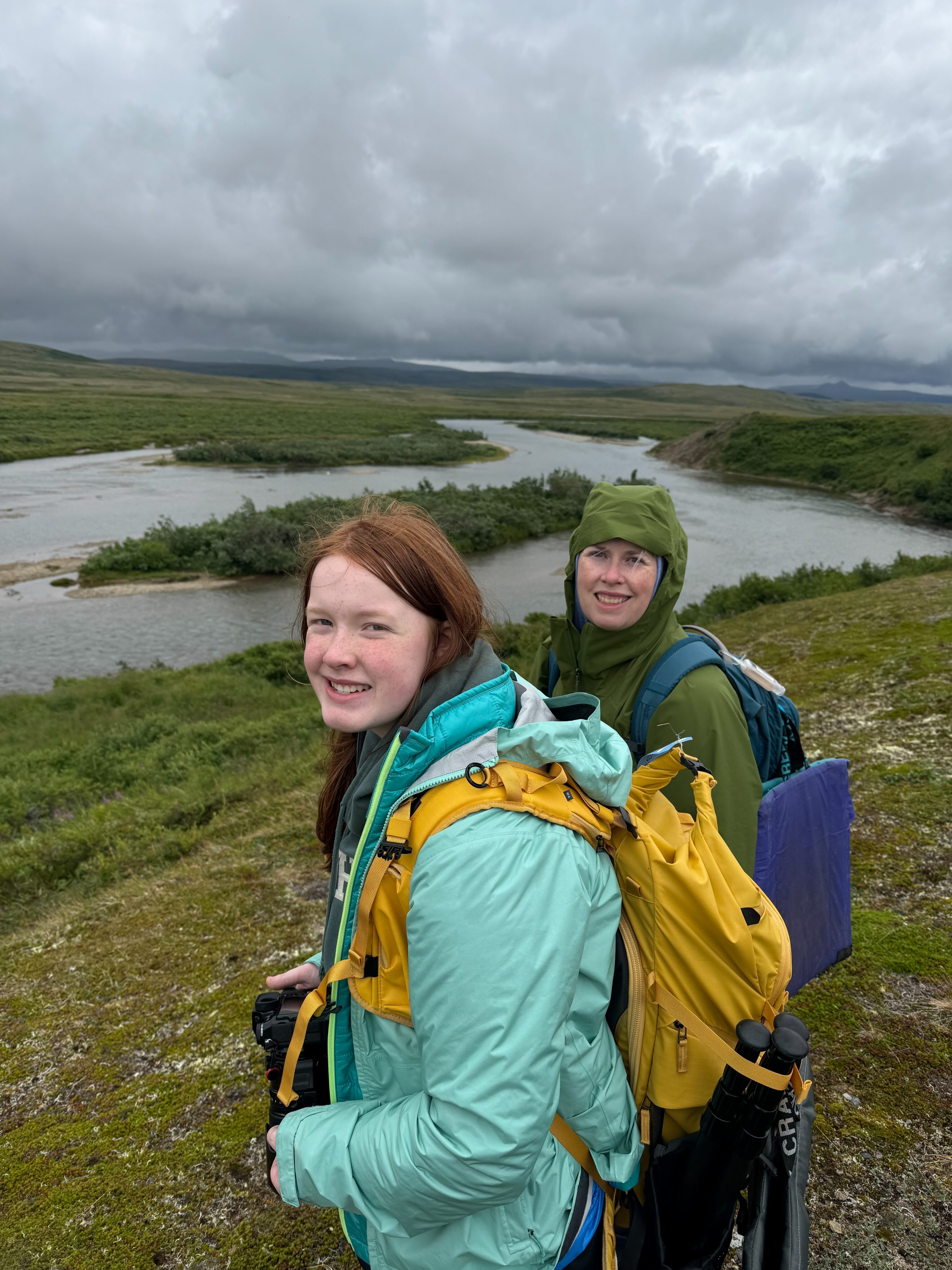 Cat and Cameron dressed in hiking gear standing in the rain on the banks of the river deep in the back country of Katmai National Park.