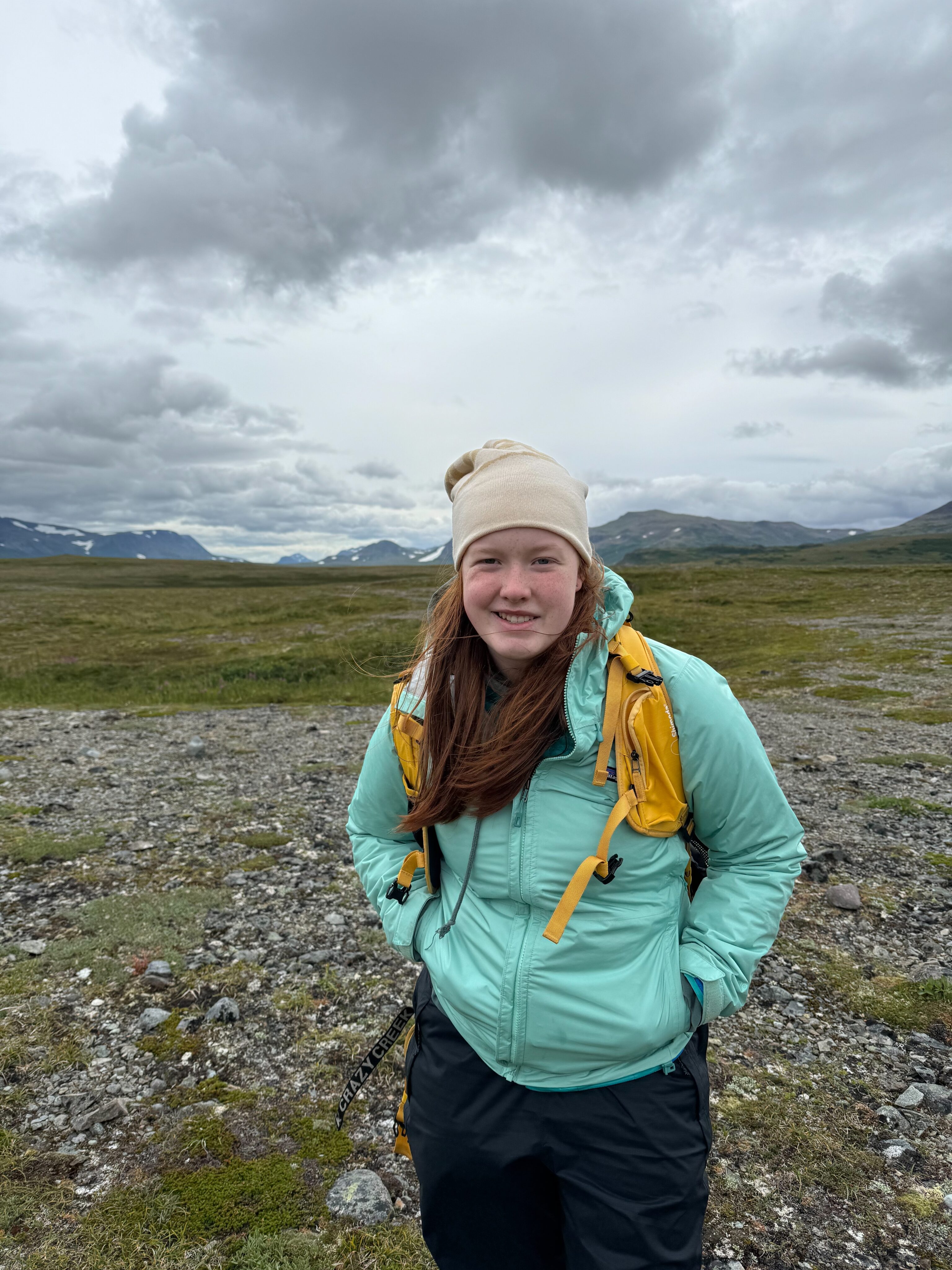 Cameron wearing a blue rain jacket, white hat and yellow camera bag, standing on the tundra on a cold day in the wilderness of Katmai National Park.