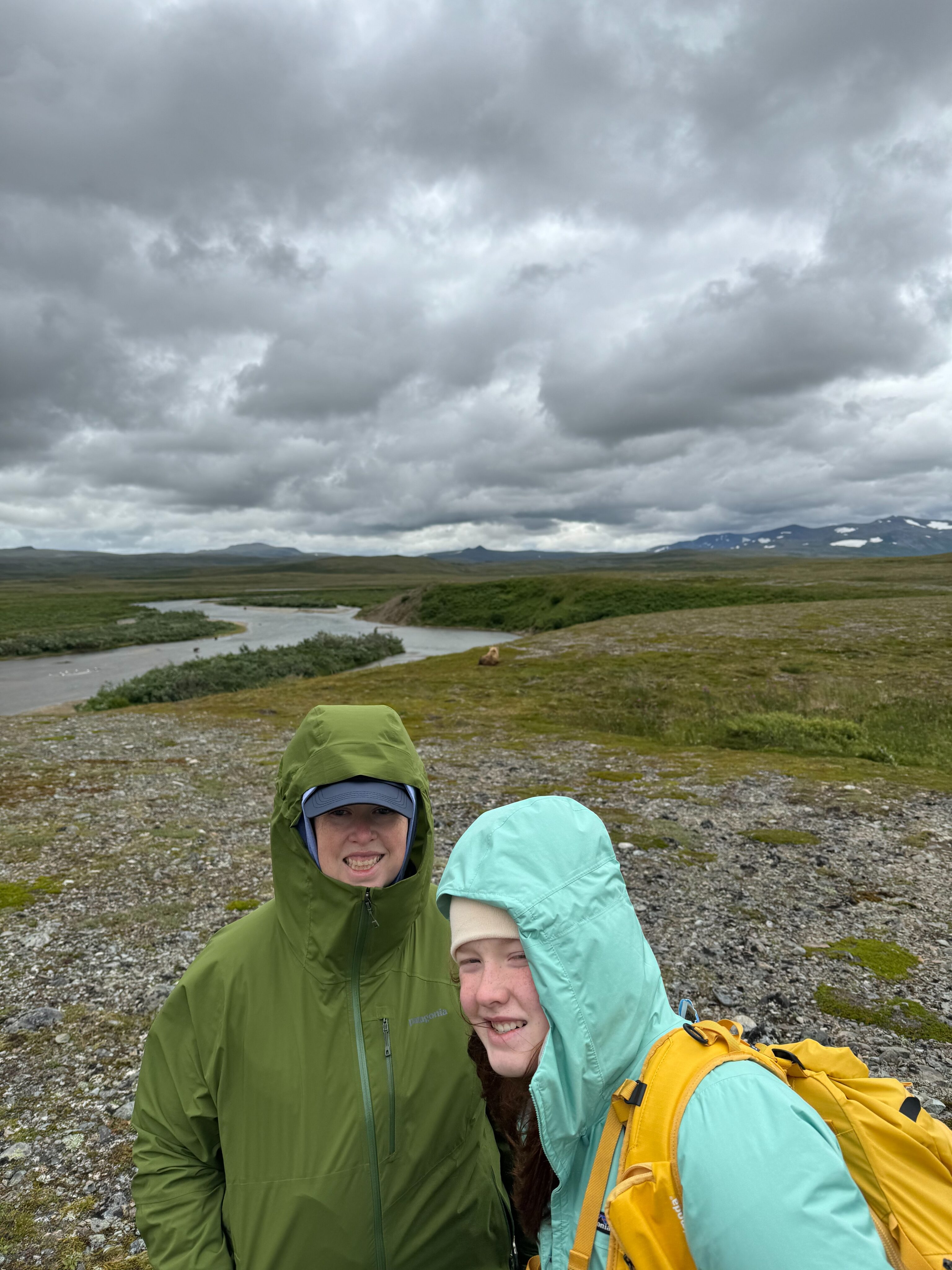 Cat and Cameron standing in the rain, both wearing full rain gear, pose for a photo high up on the banks of the river. Storm clouds are overhead. 