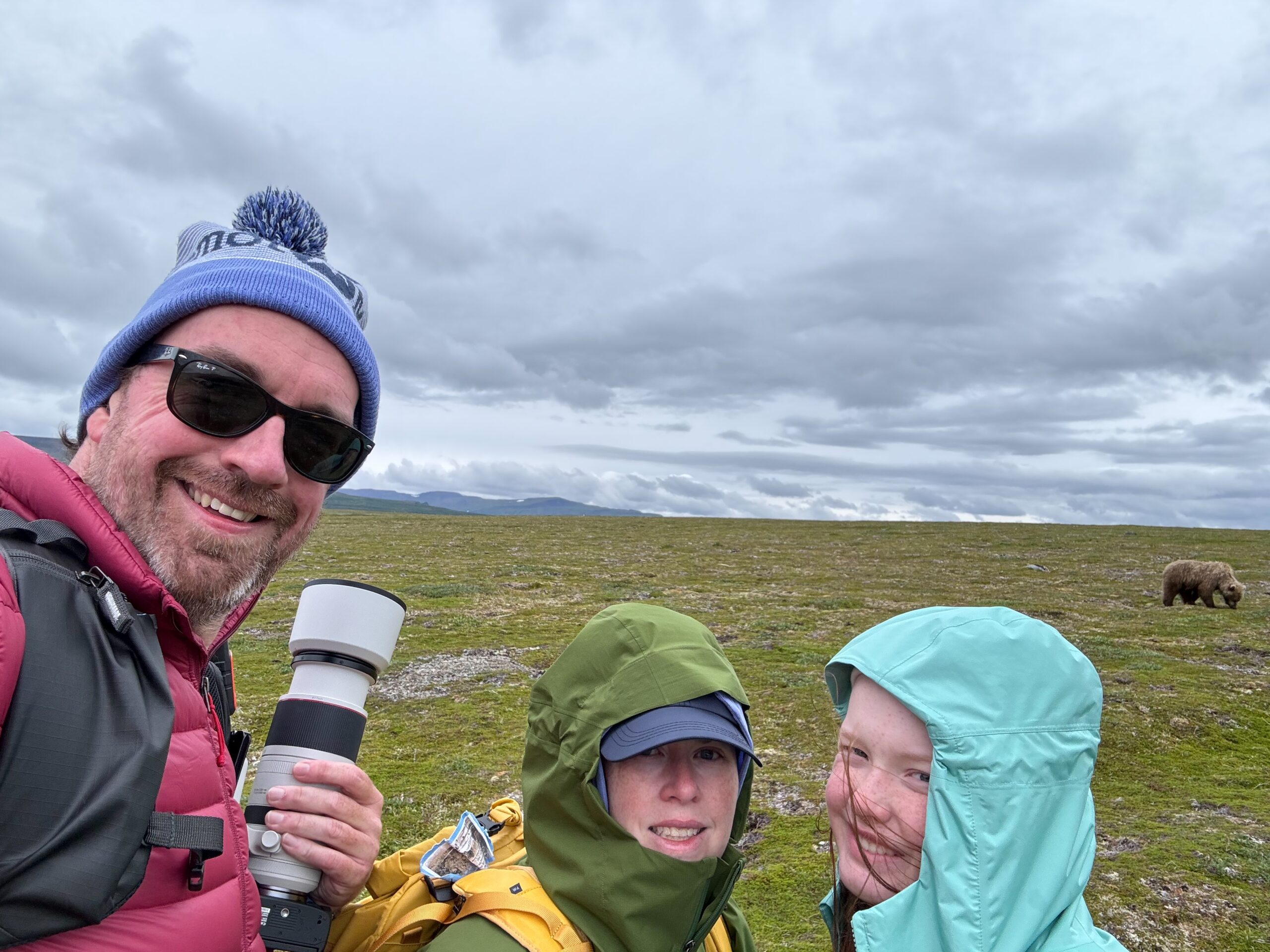 Cat, Cameron and myself pose for a selfie with a brown bear in the background. I'm wearing a hate and holding my camera while Cat and Cameron both have full raingear. 