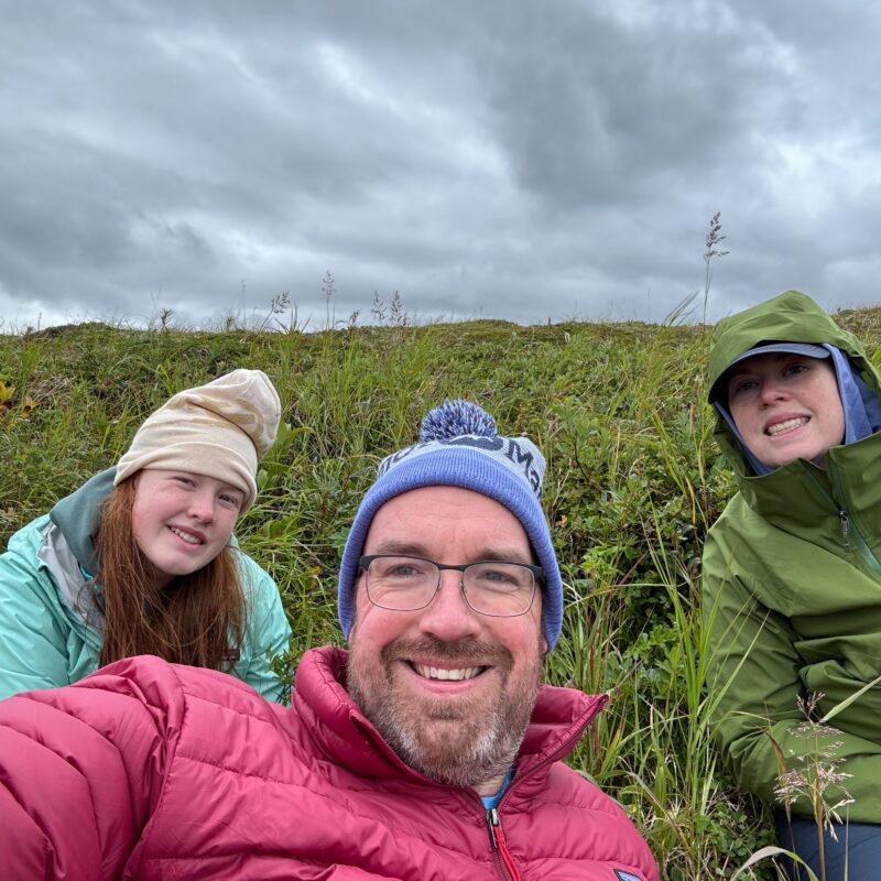 Cat, Cameron and myself all pose for a photo while sitting in the middle of the bank by the river. All of us wearing cold weather and rain gear.