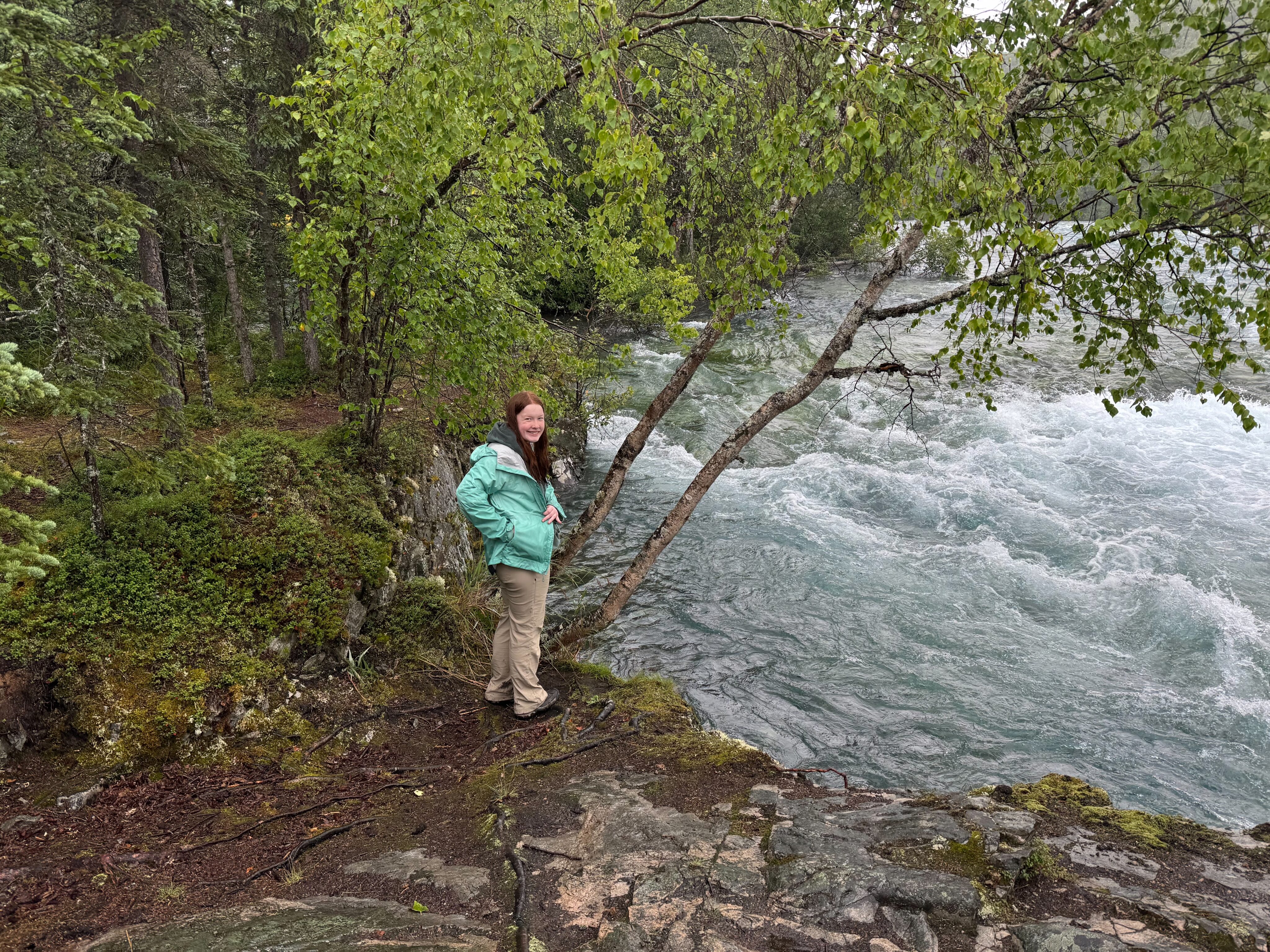 Cameron wearing a raincoat and hiking pants standing right next to Tanalian Falls in Alaska. 