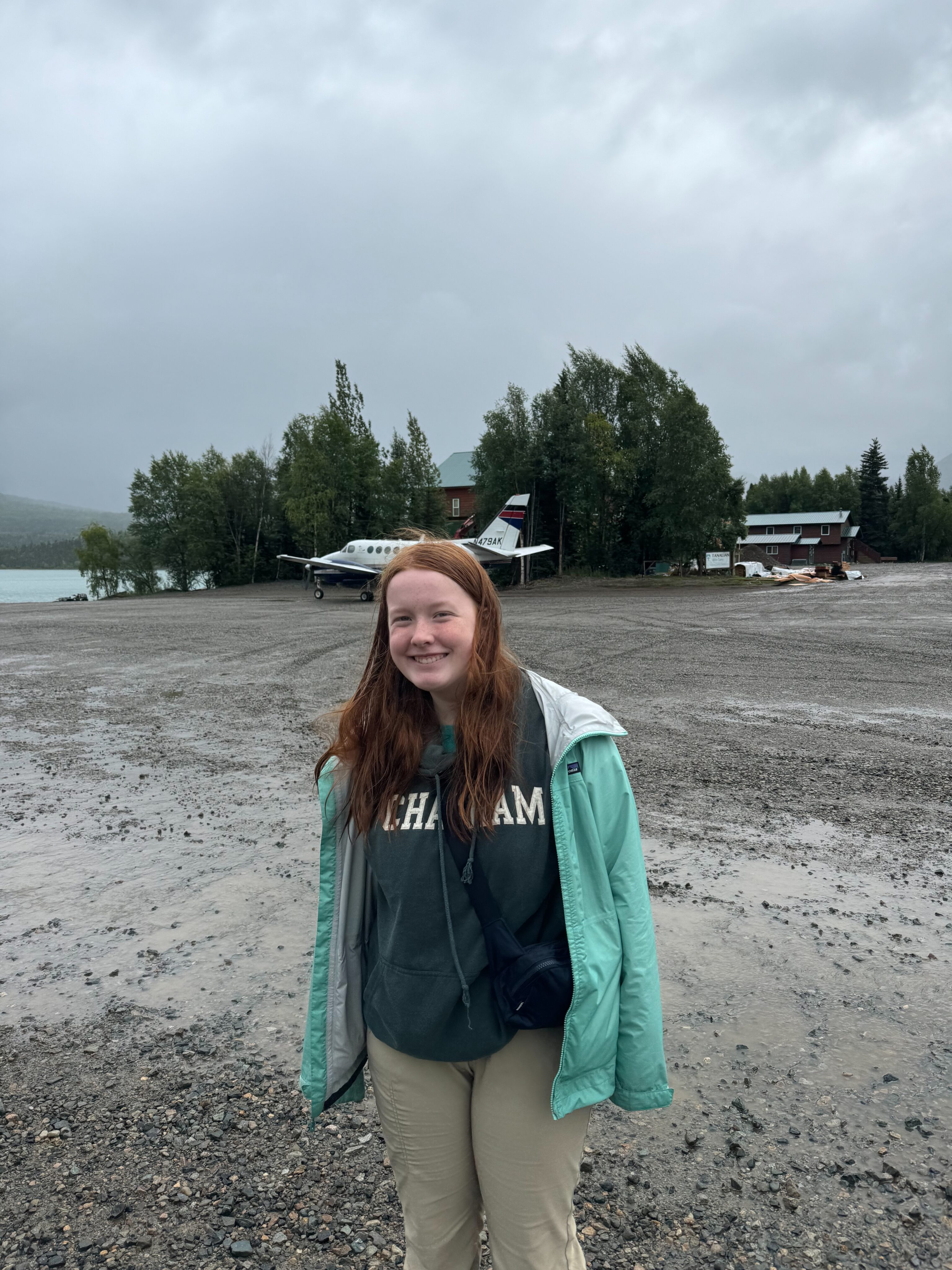 Cameron wearing a raincoat and hoodie standing on the edge of the runway in Port Alsworth.