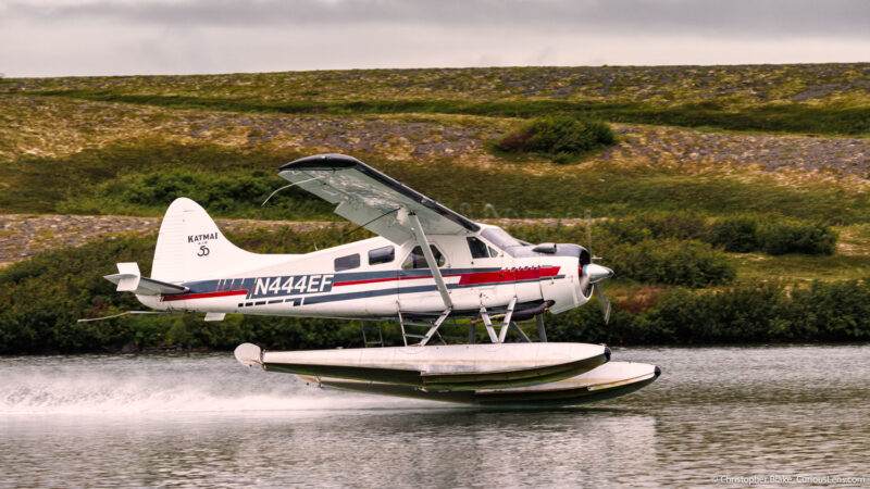 Float plane landing on a lake in Katmai National Park, Alaska, with rugged landscape in the background, capturing the adventurous spirit of the park.