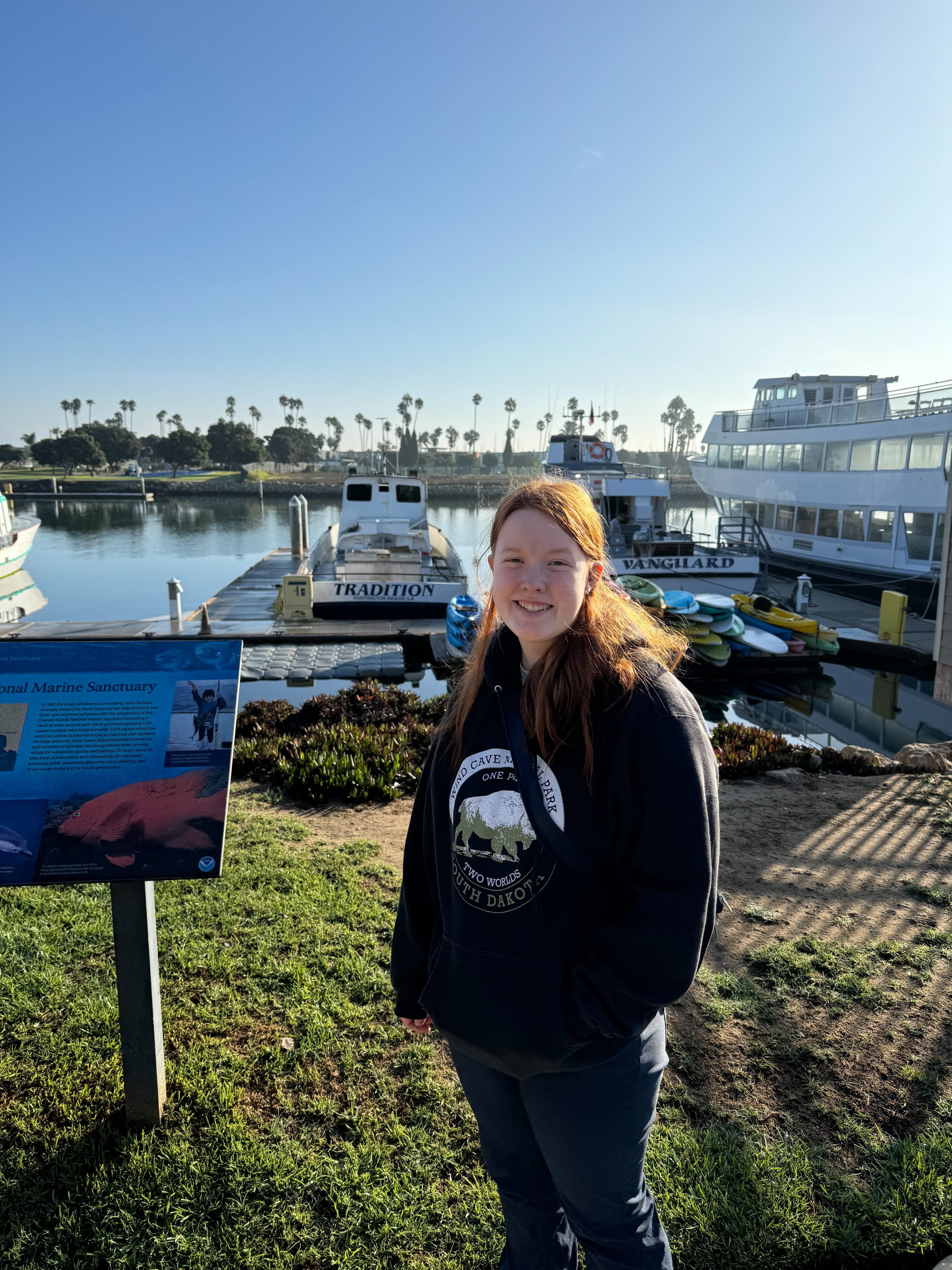 Cameron wearing a sweatshirt standing in the morning sun with a big smile on her face. Outside on the grass in the harbor waiting for the island packers boat.