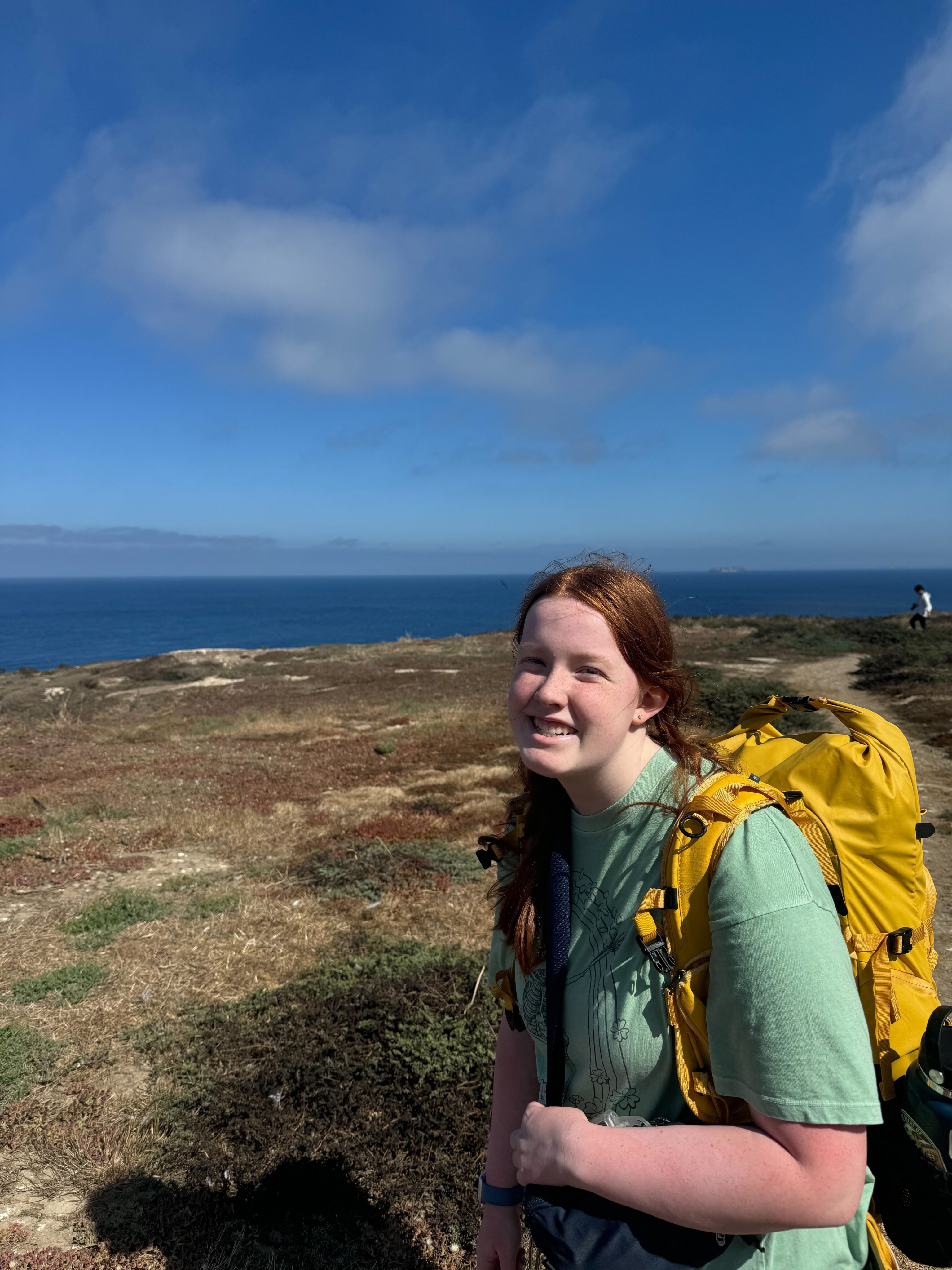 Cameron with a big smile, in a t-shirt and yellow camera bag, stopped along the hiking trail on Anacap Island with the great view of the Pacific Ocean behind her.