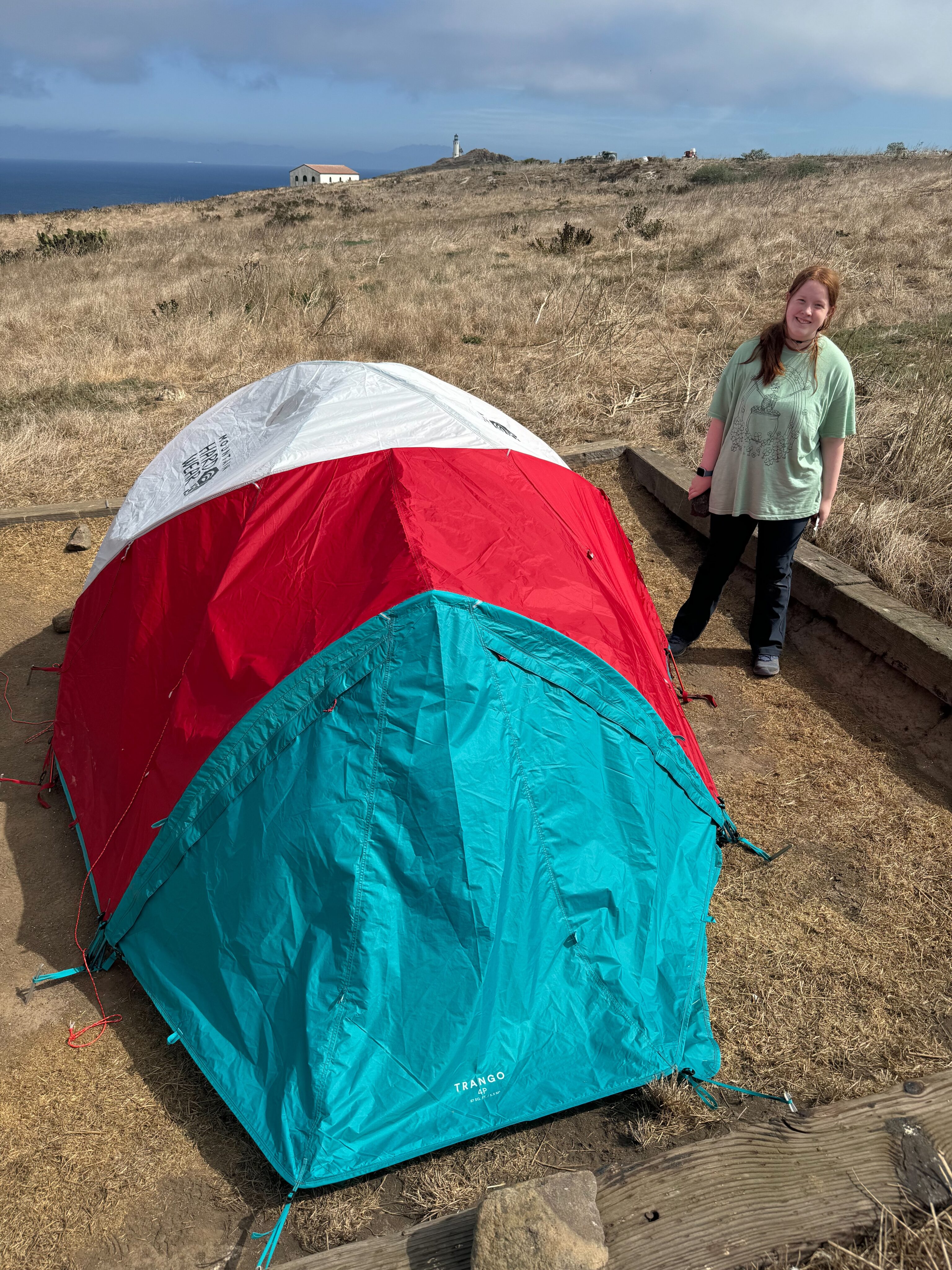 Cameron in hiking pants and a t-shirt standing next ot our text at our campsite in Anacapa with a view of the ocean behind us.