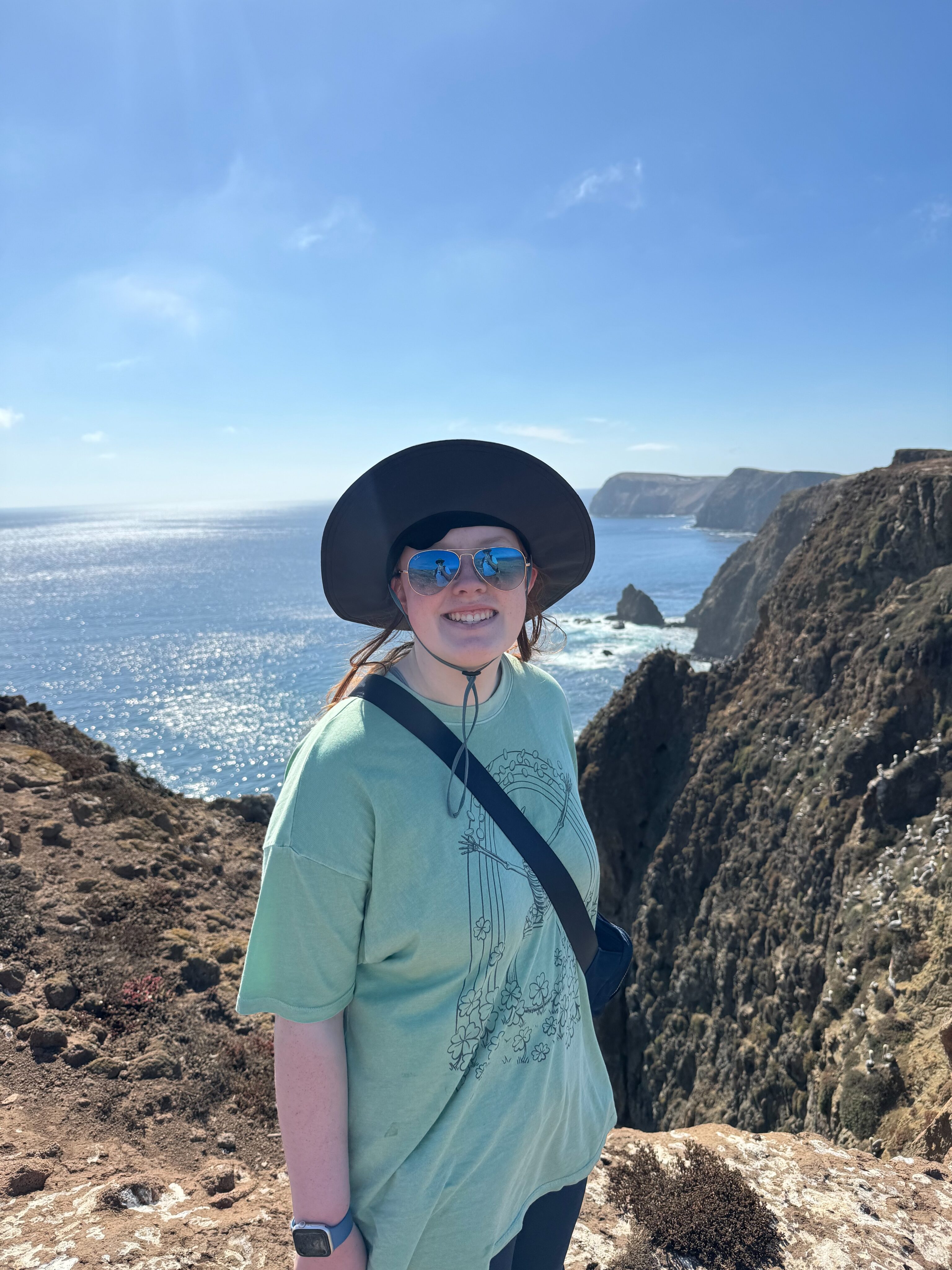 Cameron stops for a photo on a hiking trial on Anacapa Island, wearing a t-shirt, hat and sunglasses. Sharp cliffs and the ocean are in the background on a clear blue sky day.