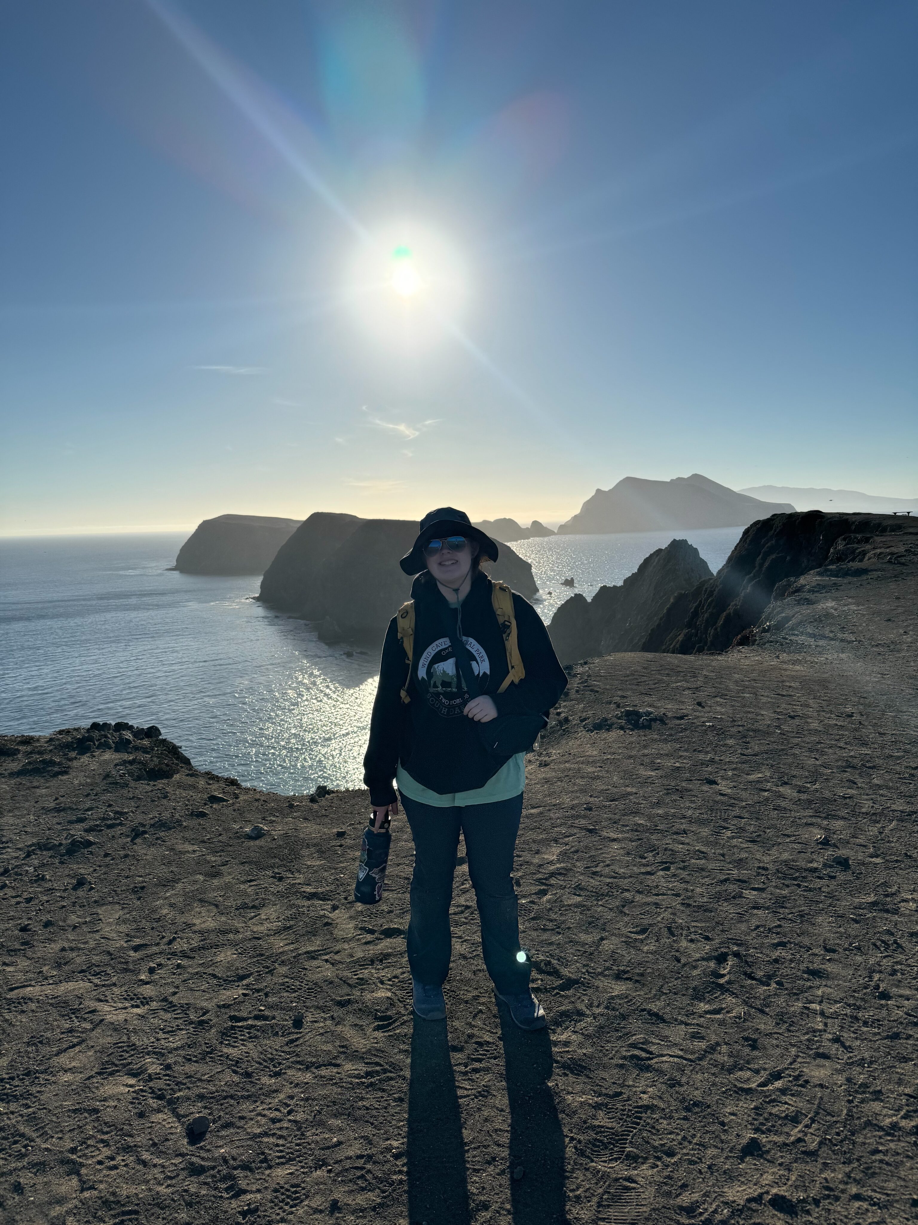 Cameron in a hoodie, hat and sunglasses standing at the end of the trial at Inspiration Point on the Channel Islands National Park. The sun is just above her and the other chain of islands is behind her.