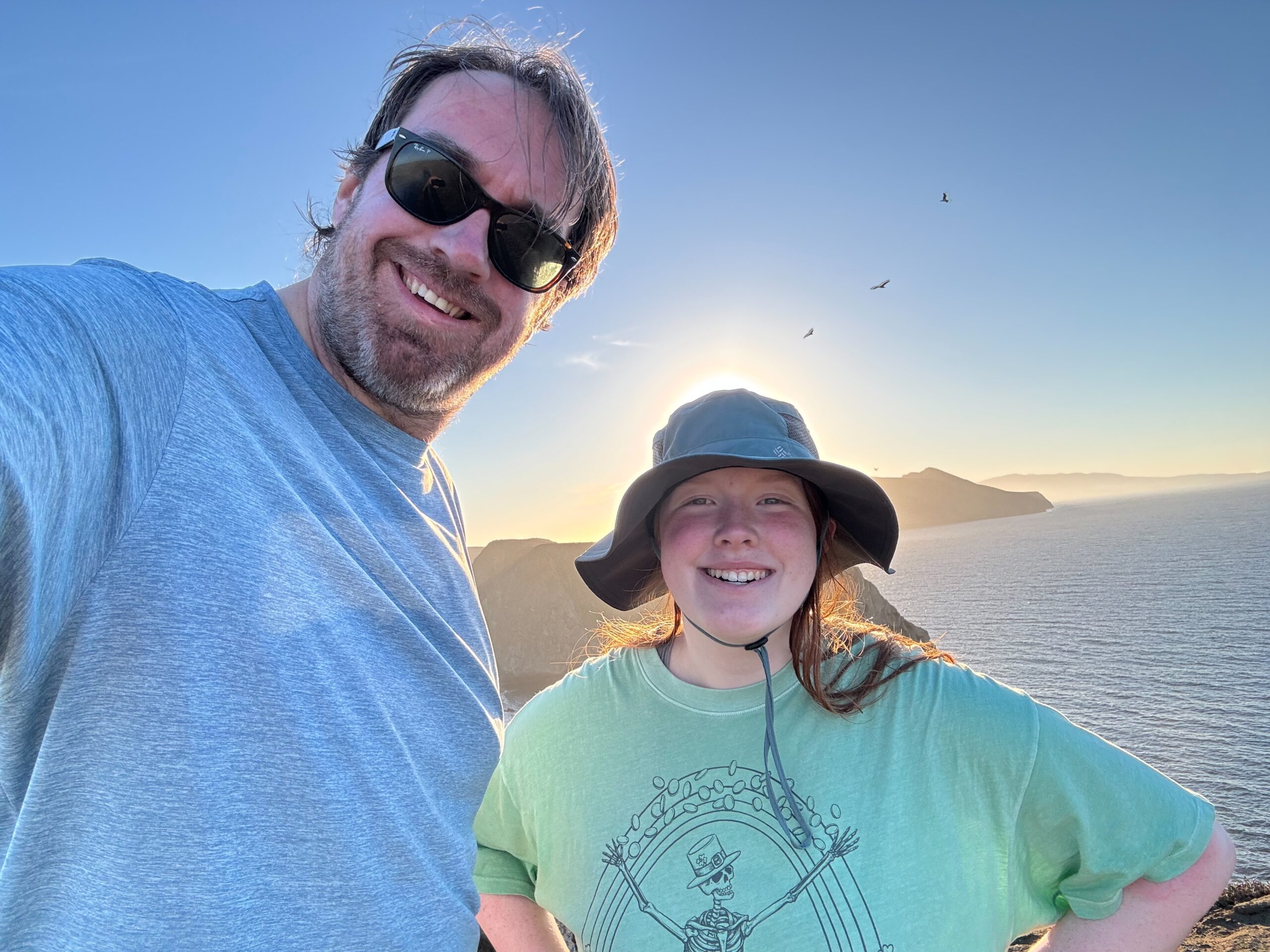 Cameron in a t-shirt and had, and myself in a t-shirt and sunglasses pose for a photo at Inspiration Point on the channel islands. The sun dis directly behind Cameron and is creating a glow around her.