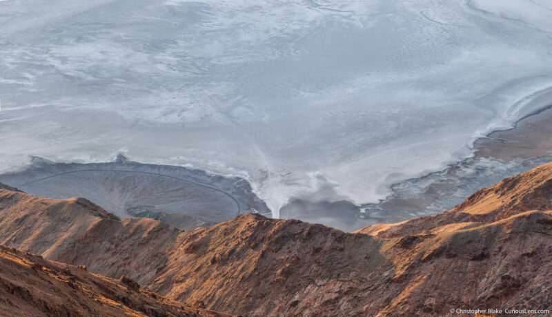 Aerial view from Dante's View showing the last sunlight on the rocky foreground with Badwater Basin's salt flats and a winding road in the distance, capturing the vastness of Death Valley.