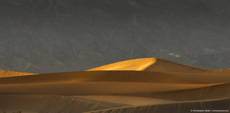 Golden light on a large sand dune at Mesquite Sand Dunes, with smaller dunes in the foreground and shadowed mountains in the background during sunset.