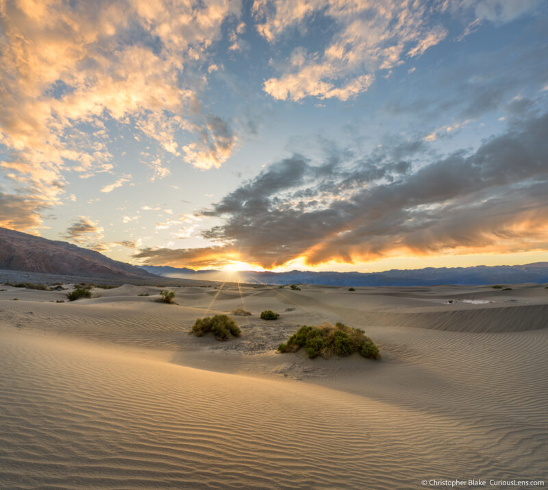 Late November sunset at Mesquite Flat Sand Dunes featuring a sunstar over the mountains, with foreground of desert brush and sand patterns leading into the dramatic sky.