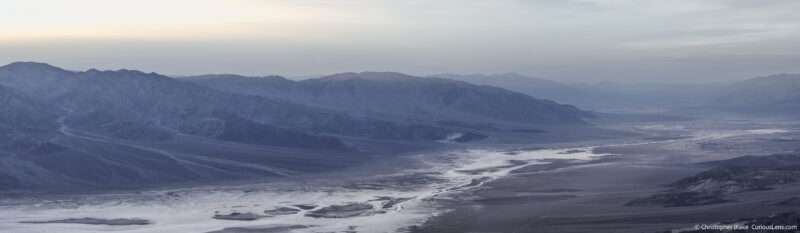 Panoramic view from Dante's View at dusk showing the vast Badwater Basin and salt flats leading into the layered mountain ranges, with a haze enhancing the depth and scale of the scene.