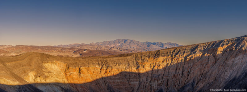 Panoramic view of Ubehebe Crater at sunset, highlighting the detailed textures of the crater rim with a dramatic shadow line and distant mountain ranges in Death Valley National Park.