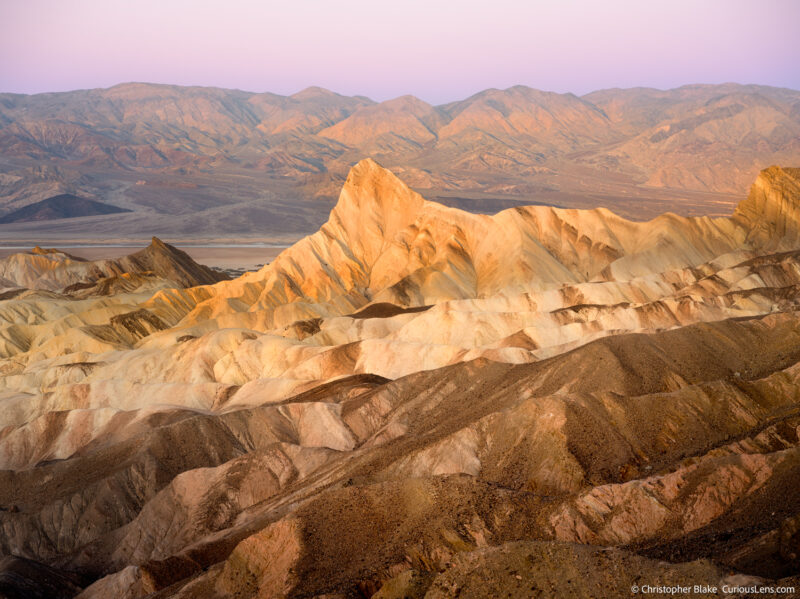 Early morning view from Zabriskie Point with the first sunlight illuminating the Panamint Range and the badlands, under a warm purple sky.